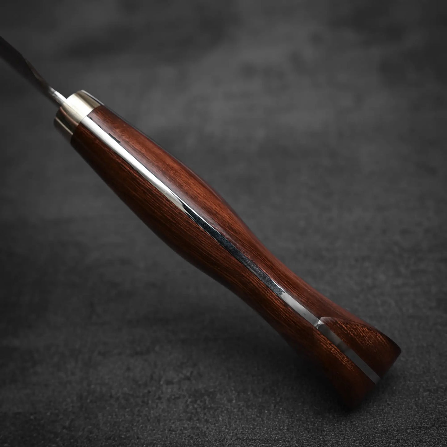 A close-up of the Shigeki Tanaka R2 Damascus Gyuto 270mm - Ironwood shows its polished dark wooden handle with metal collar on a textured surface, highlighting the smooth contours and glossy finish that complement its damascus cladding.