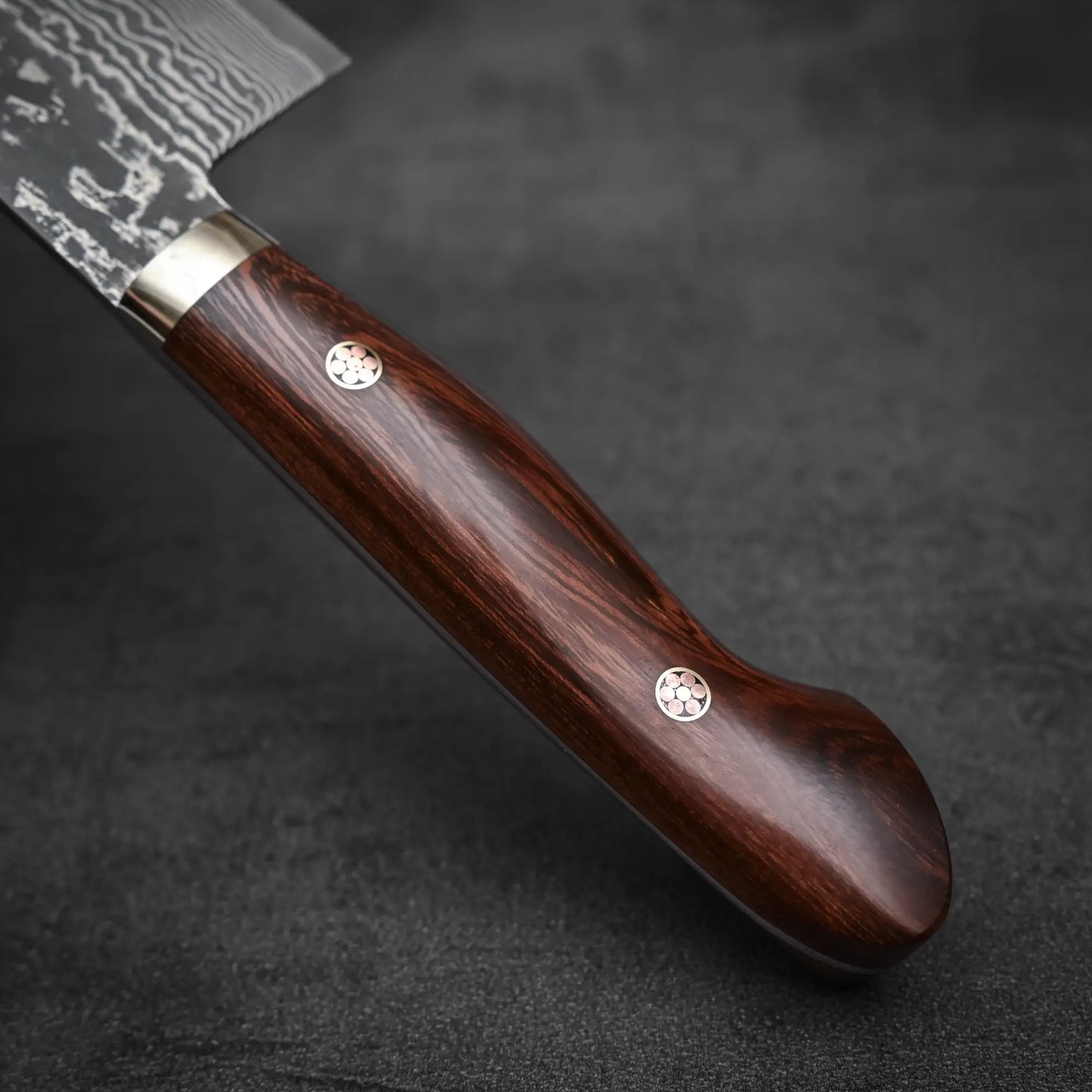 Close-up of the Shigeki Tanaka R2 damascus gyuto 270mm - ironwood handle, crafted from dark polished wood with two metal rivets. The blade displays a distinctive wavy Damascus pattern near the handle, set against a dark background.
