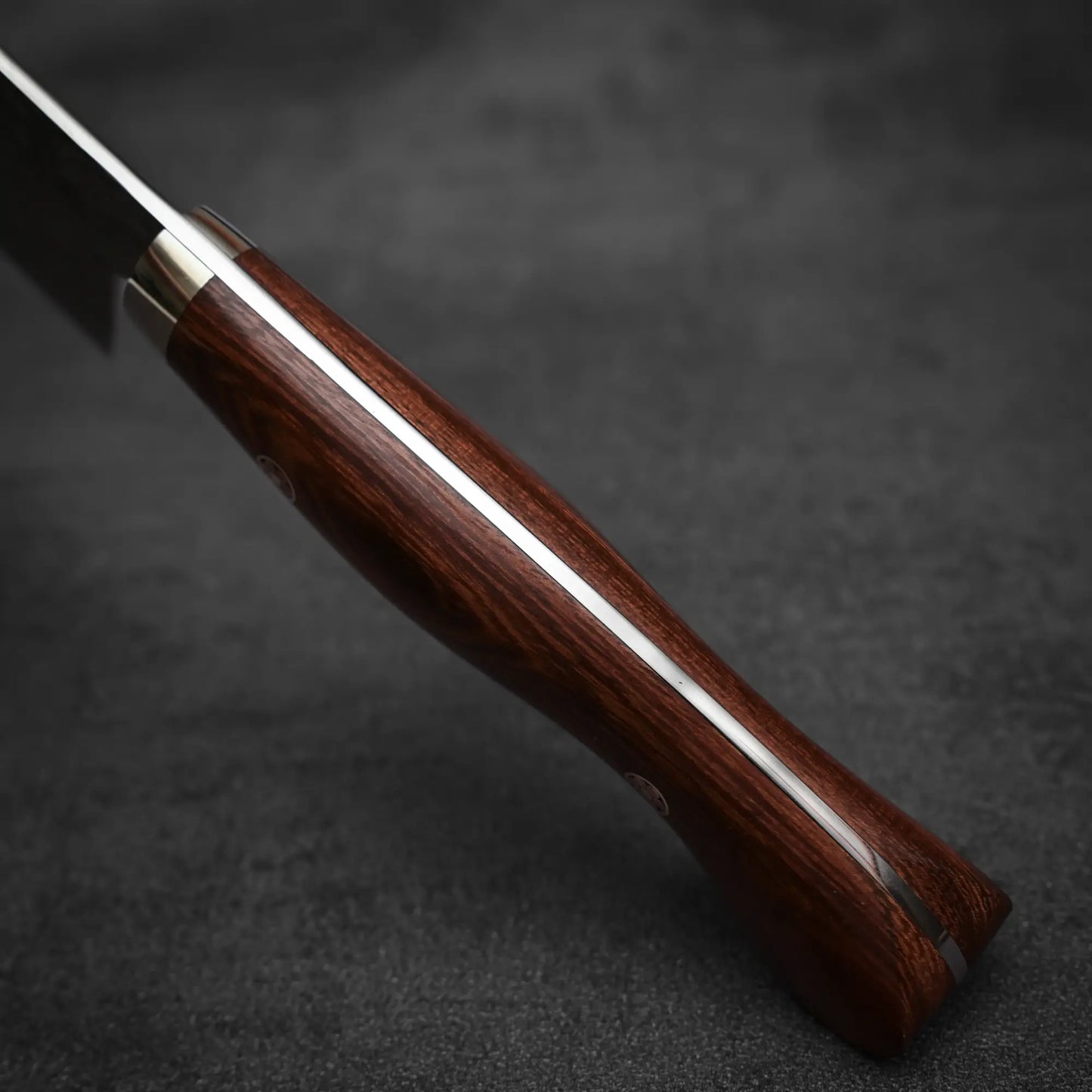 Close-up of the handle on a Shigeki Tanaka R2 damascus gyuto 270mm - ironwood, showing its dark polished ironwood grip and exposed metal tang, resting on a dark textured surface.