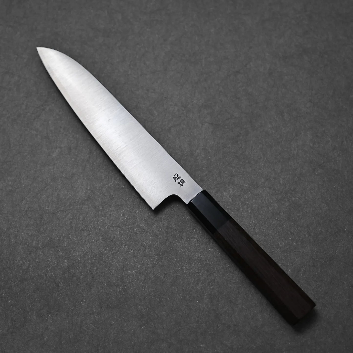 The Sukenari ZDP189 gyuto 195mm, featuring a silver ZDP189 stainless steel blade and dark wooden handle, is placed on a textured gray surface.