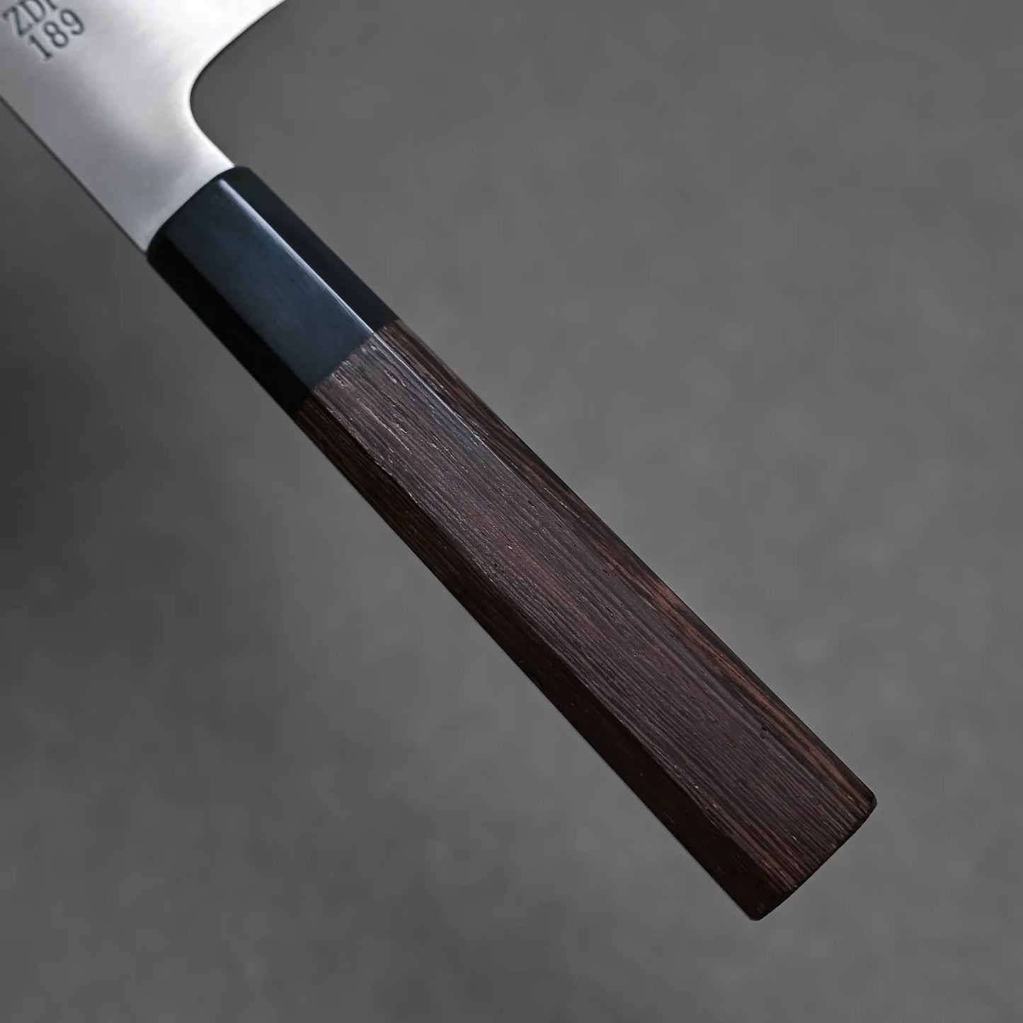 Close-up of the Sukenari ZDP189 gyuto 195mm handle made of dark wood with a black bolster on a gray background. The Sukenari blade in ZDP189 stainless steel is partially visible at the top left.