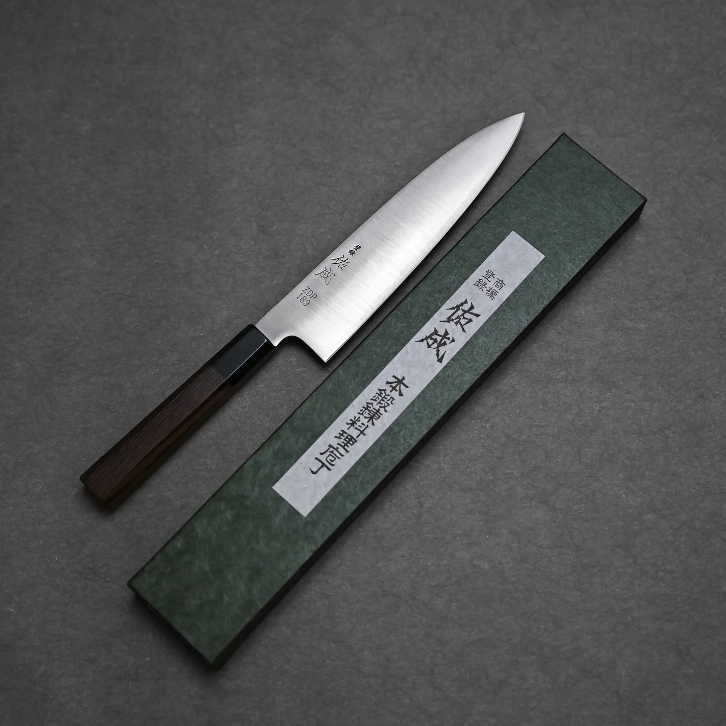 The Sukenari ZDP189 gyuto 210mm chefs knife with a wooden handle rests beside a green rectangular box with Japanese text on white labels, all set on a dark gray surface.