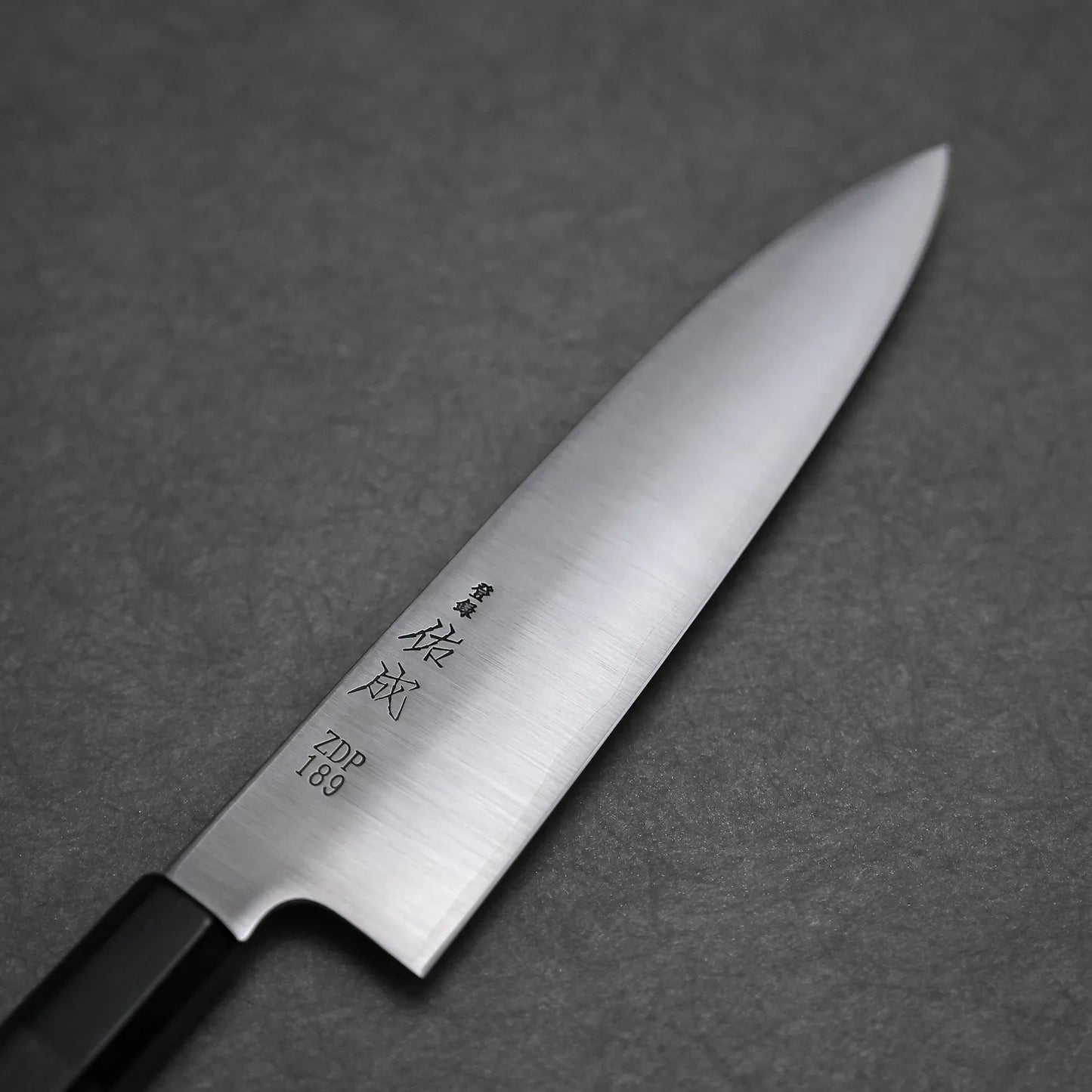 A close-up of the Sukenari ZDP189 gyuto 210mm, a Japanese kitchen knife by Sukenari with engraved kanji on its ZDP189 stainless steel blade, resting on a dark gray textured surface. Handcrafted in Toyama, Japan.