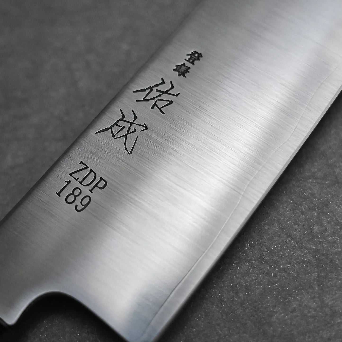 A close-up of the Sukenari ZDP189 gyuto 210mm shows its ZDP189 steel blade with engraved Japanese characters and a brushed metal finish, set against a dark, textured background that reflects its Toyama, Japan heritage.