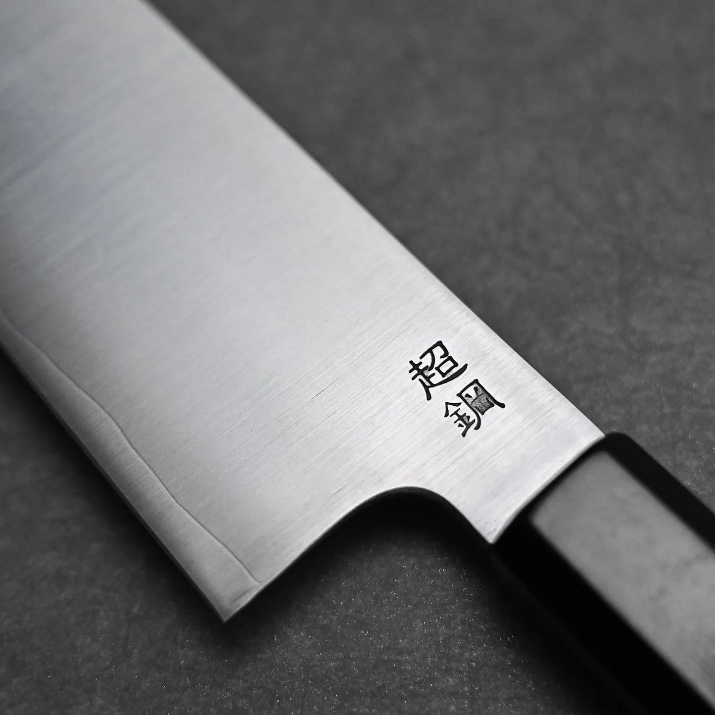 Close-up of the Sukenari ZDP189 gyuto 210mm kitchen knife, crafted from premium stainless steel with kanji characters engraved on the blade, by Sukenari, displayed on a dark textured surface.