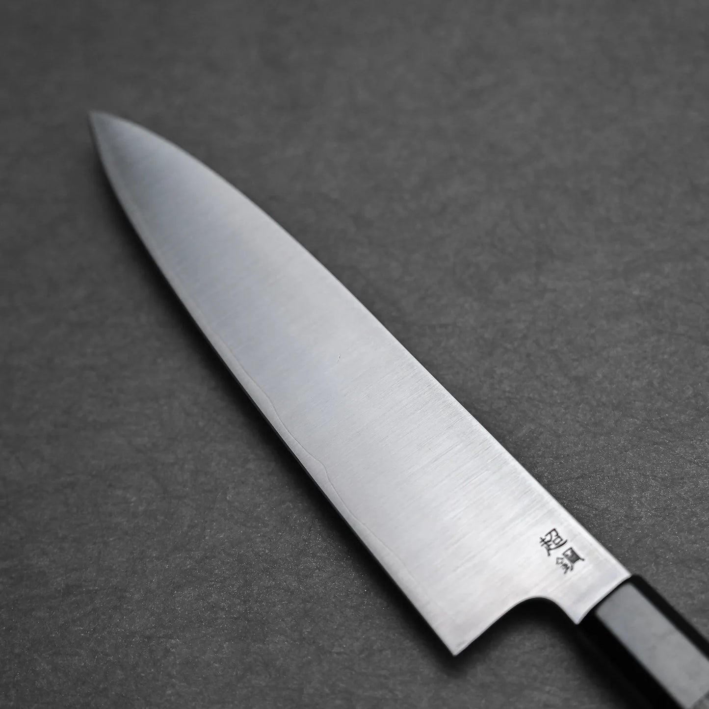 A close-up of the Sukenari ZDP189 gyuto 210mm chef’s knife with a black handle rests on a dark, textured surface. The sharp blade, by Sukenari, features Japanese characters engraved near the base.