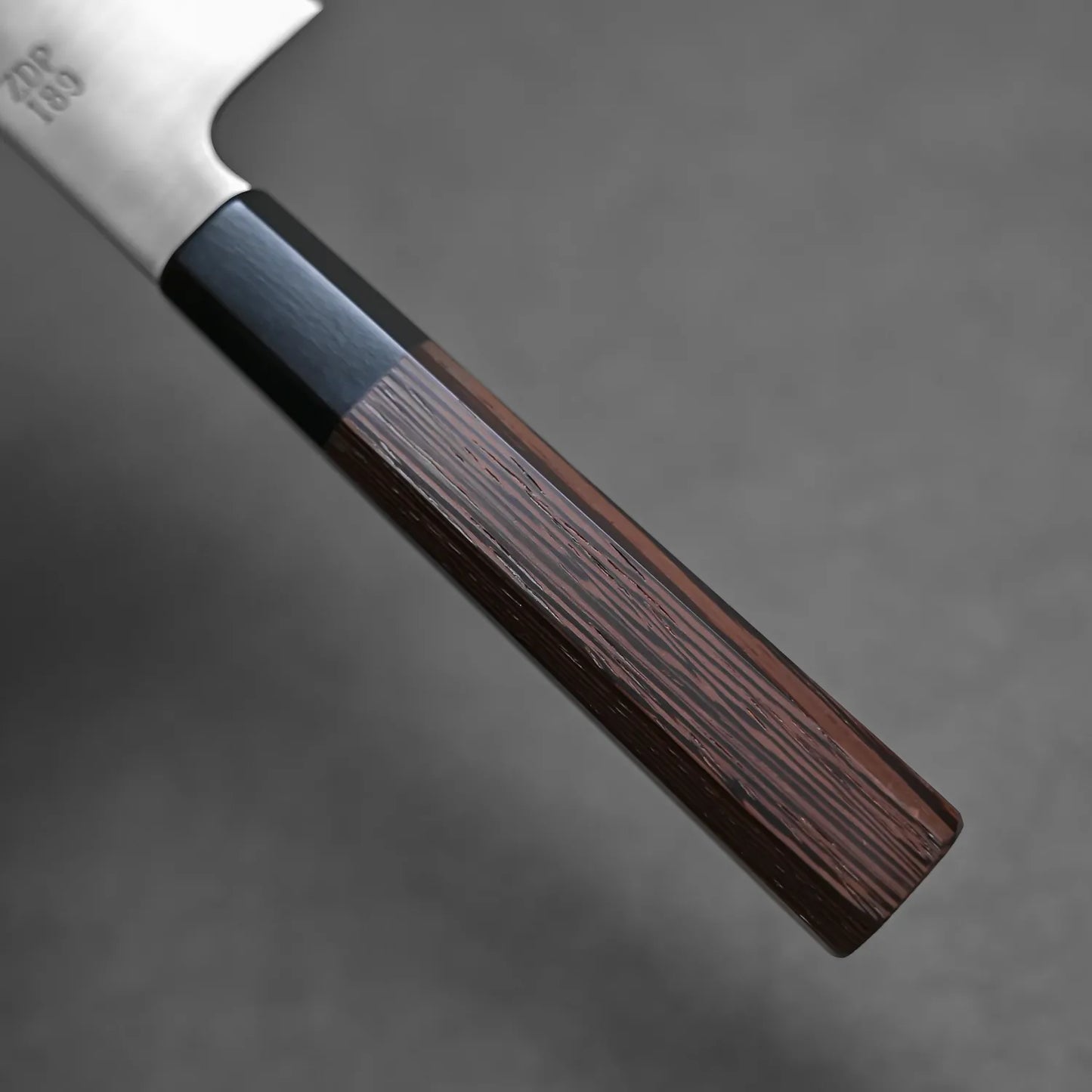 Close-up of the Sukenari ZDP189 gyuto 210mm handle, featuring two-tone dark brown wood and a black segment near the blade, set against a gray background. Crafted in Toyama, Japan by Sukenari, this knife highlights exceptional craftsmanship.