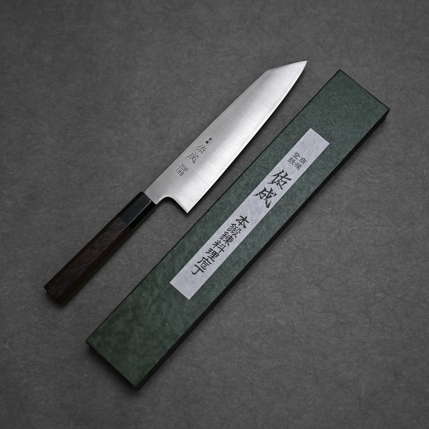 The Sukenari ZDP189 kiritsuke gyuto 240mm, a Japanese kitchen knife with a dark wooden handle, crafted by Sukenari in Toyama, Japan, lies beside a green box with Japanese text on a white label, on a gray surface.