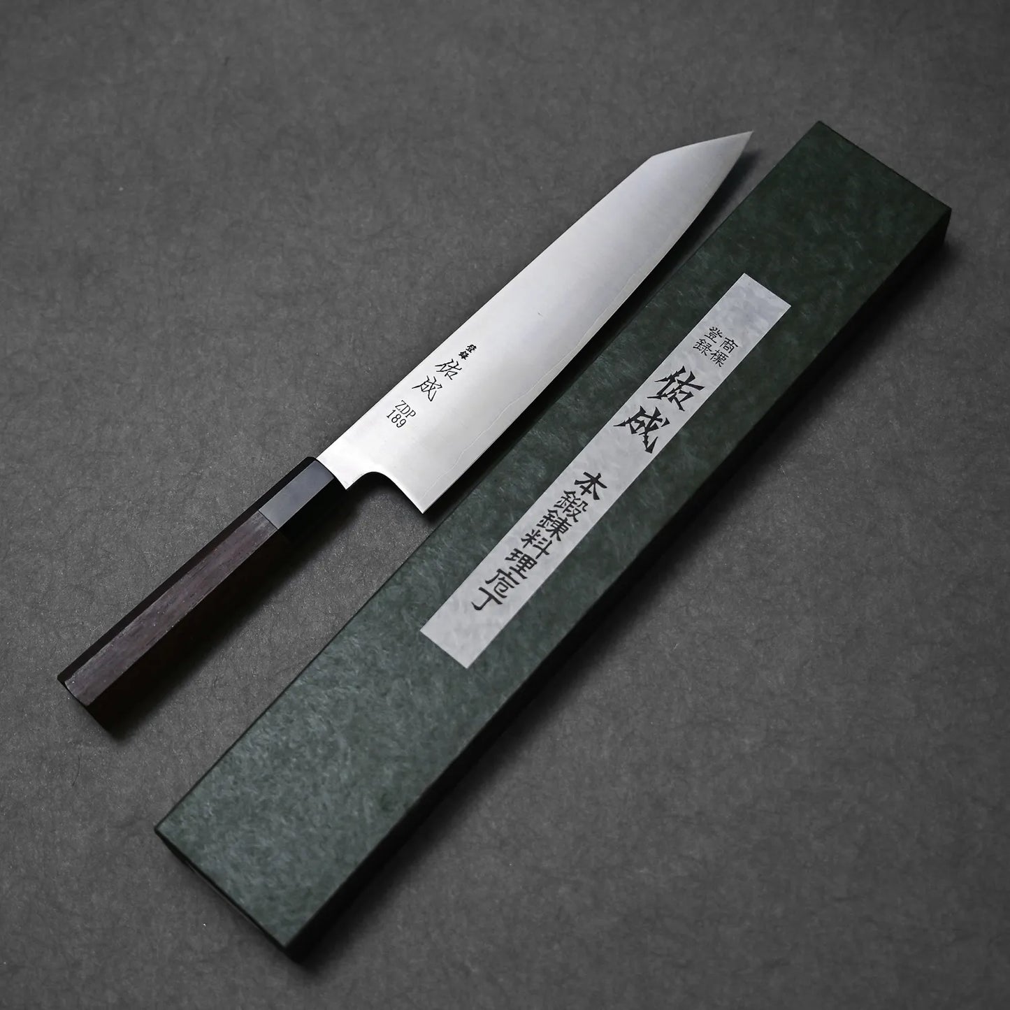 The Sukenari ZDP189 kiritsuke gyuto 270mm, a Japanese kitchen knife with exceptional edge retention and a wooden handle, is shown next to a green rectangular box with Japanese characters on a gray surface.
