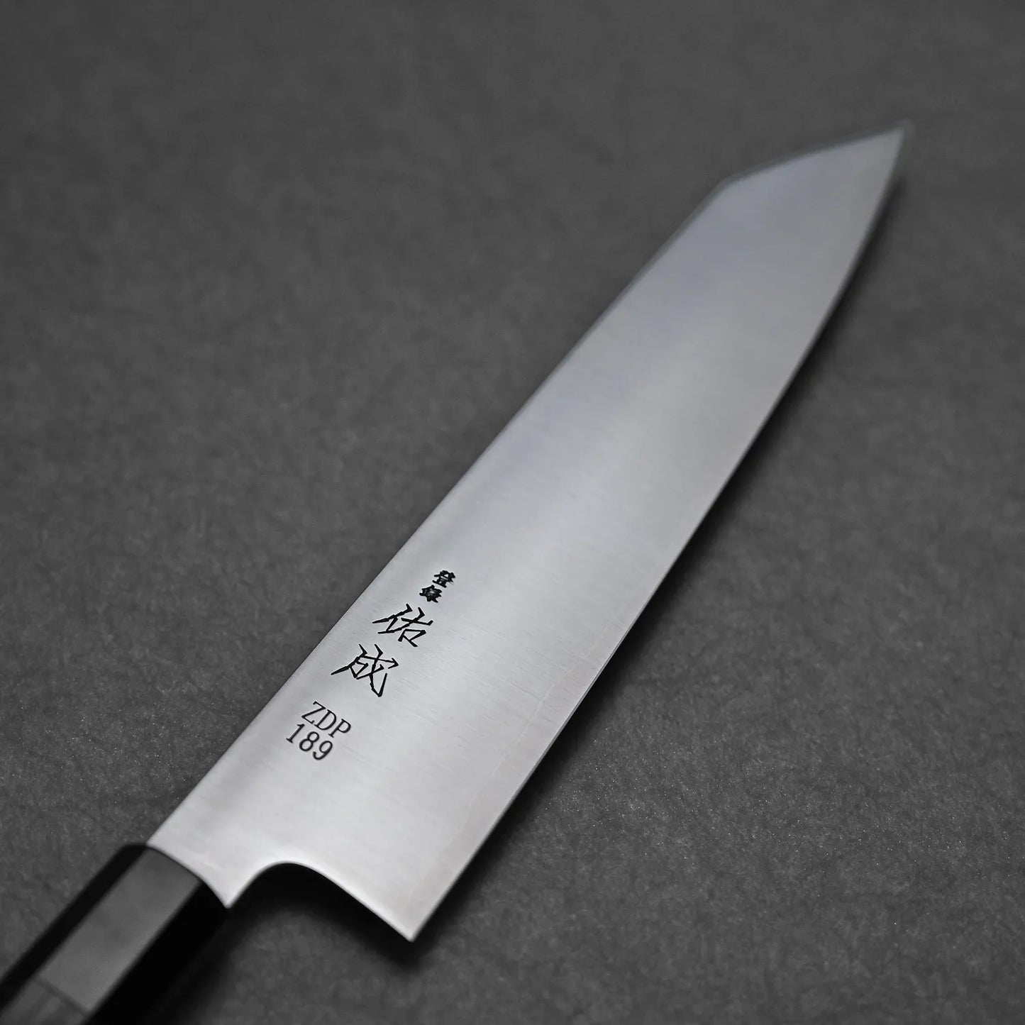 A close-up of the Sukenari ZDP189 kiritsuke gyuto 270mm chefs knife, featuring a polished blade with Japanese engravings and premium ZDP189 stainless steel near the handle, renowned for superior edge retention, displayed on a dark textured surface.