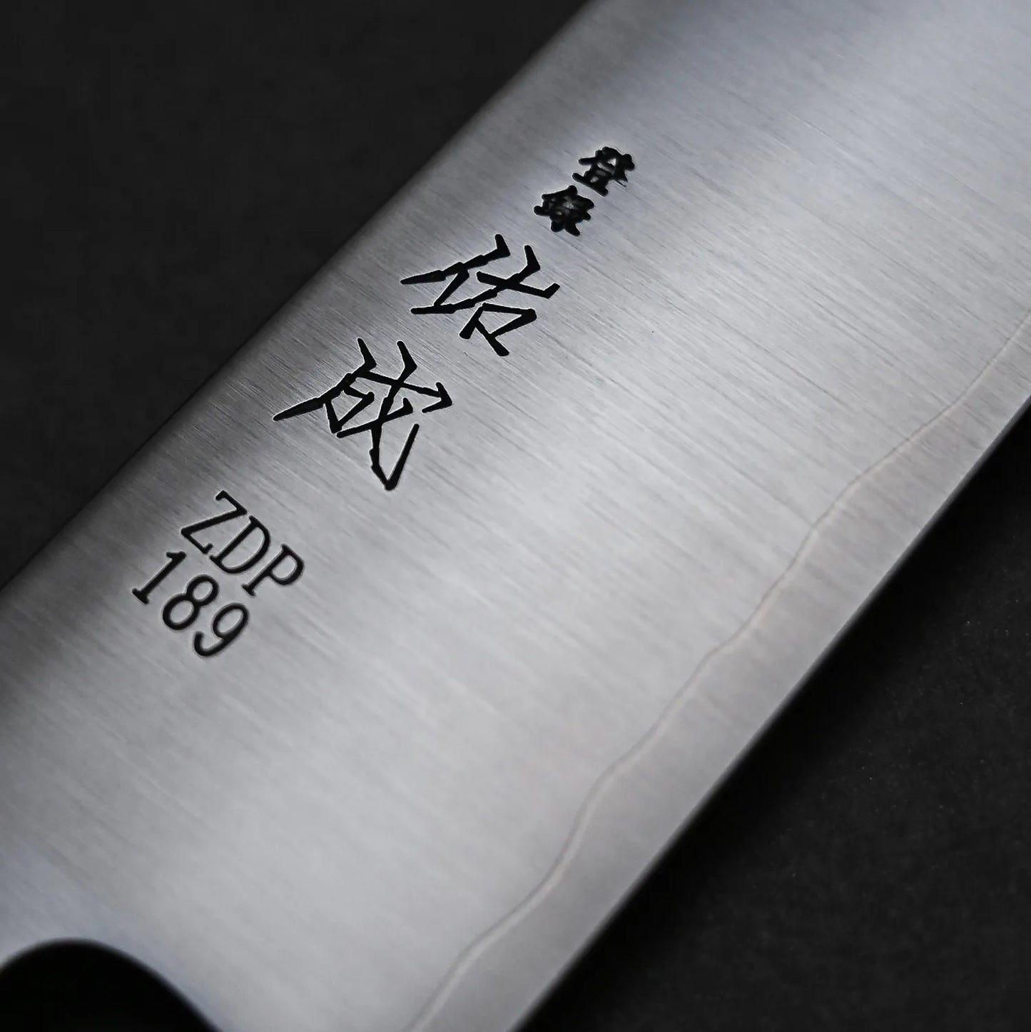 A close-up of the Sukenari ZDP189 kiritsuke gyuto 270mm blade shows Japanese characters and ZDP189 stainless steel engraving. The brushed finish and visible hamon line highlight Sukenaris exceptional edge retention against a black background.