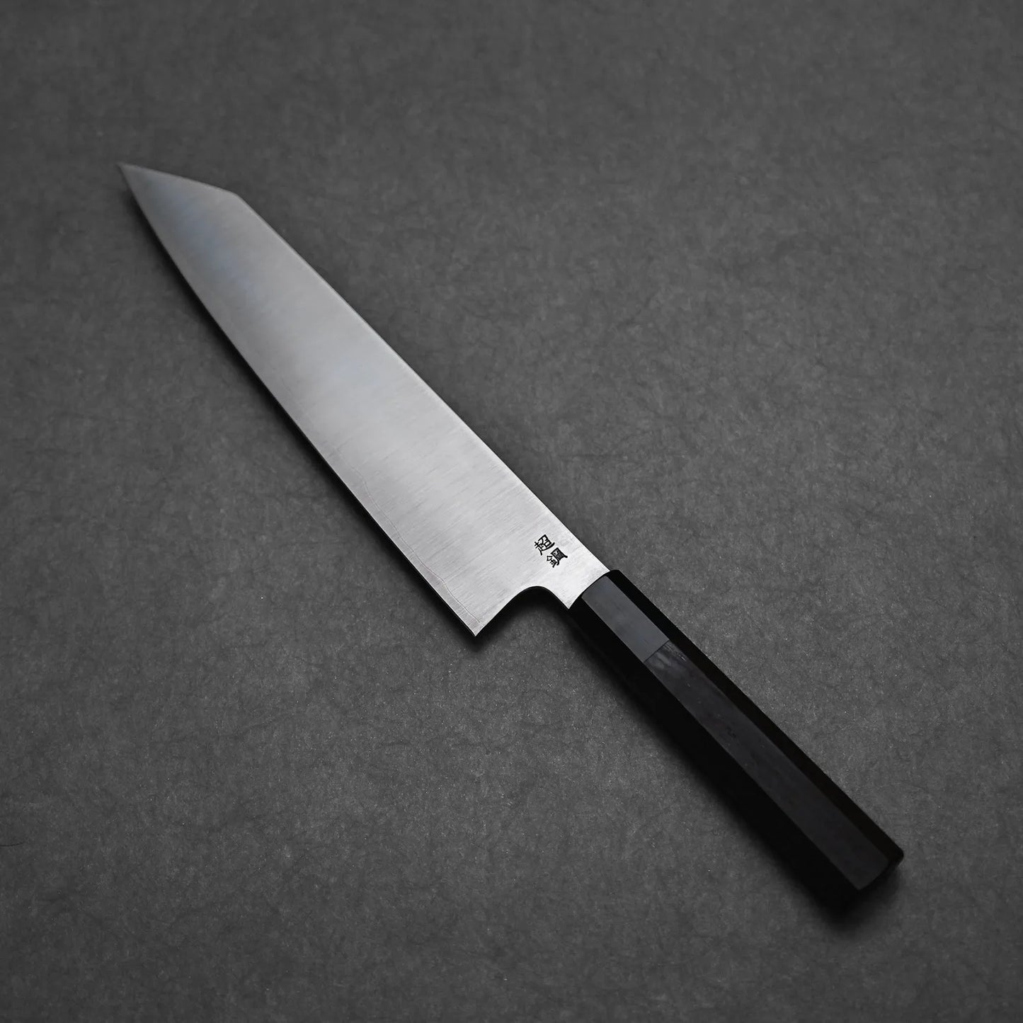 The Sukenari ZDP189 kiritsuke gyuto 270mm kitchen knife, with a sharp pointed blade and dark angular handle, rests on a textured gray surface, showcasing Sukenaris famed edge retention.