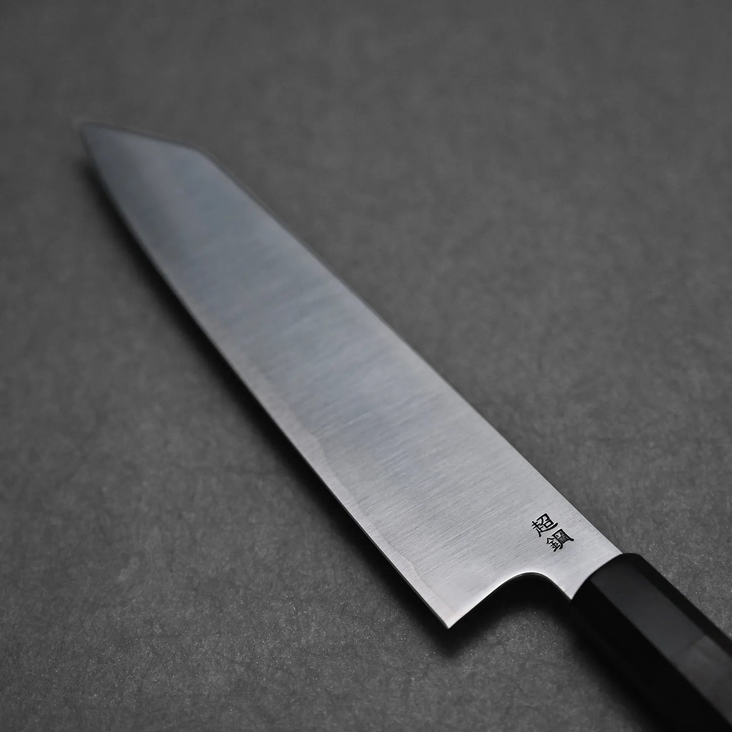 A close-up of the Sukenari ZDP189 kiritsuke gyuto 270mm shows its sharp, shiny stainless steel blade with Japanese characters engraved near the base and a black handle, set against a dark, textured surface.