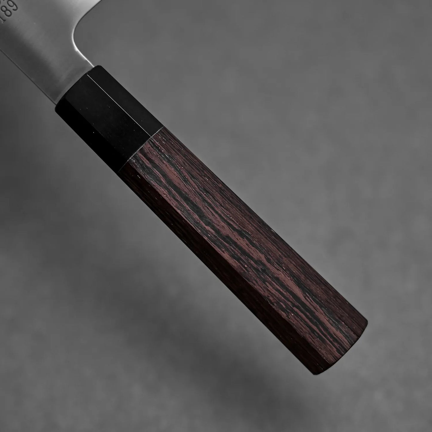 Close-up of the Sukenari ZDP189 kiritsuke gyuto 270mm: its dark wood handle with visible grain is fitted to a ZDP189 stainless steel blade renowned for edge retention, shown resting on a plain gray surface.