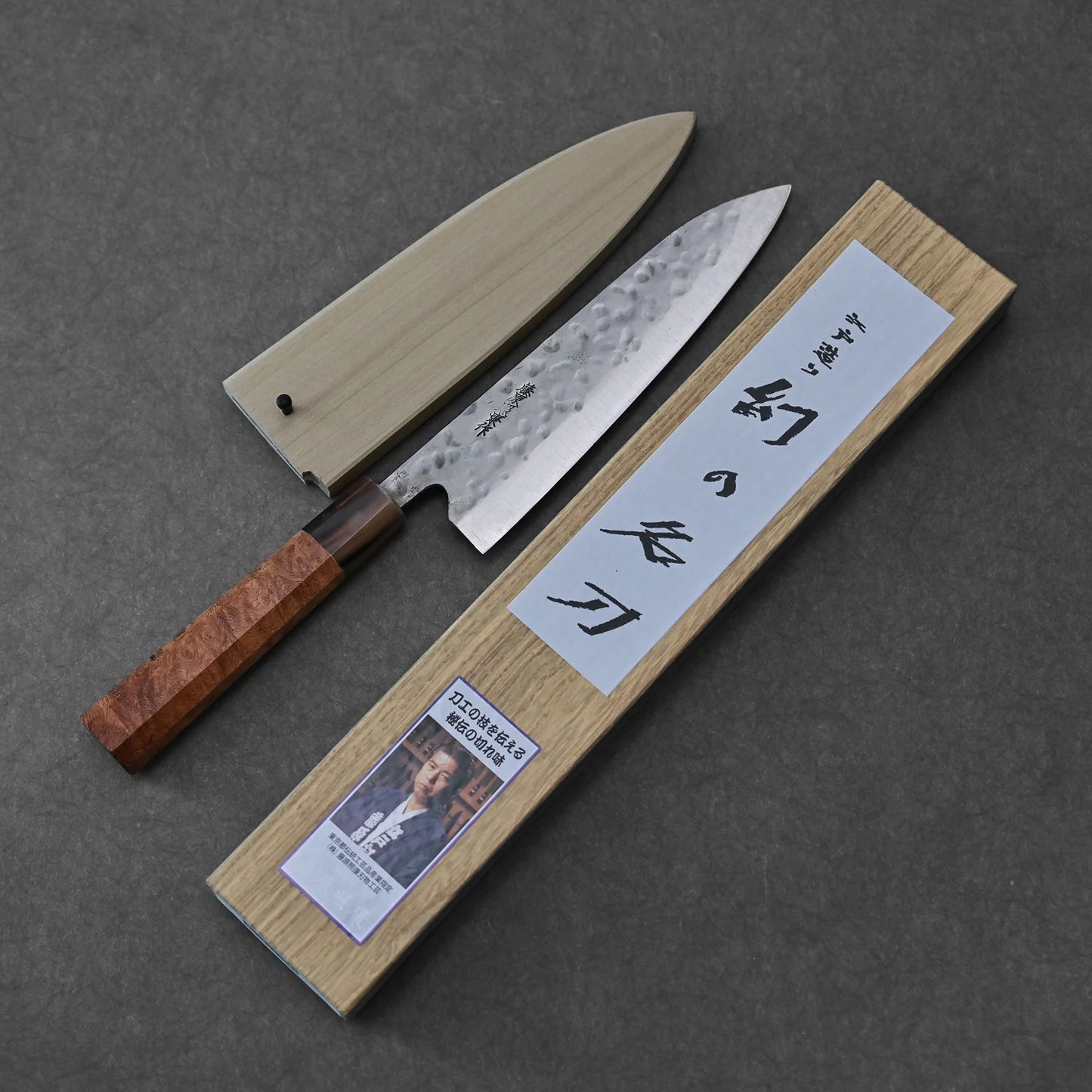 A Teruyasu Fujiwara Maboroshi shirogami#1 wa-gyuto 210mm with custom handle, saya, and polished choil by Teruyasu Fujiwara is displayed on a dark surface, accompanied by a calligraphy-adorned box.