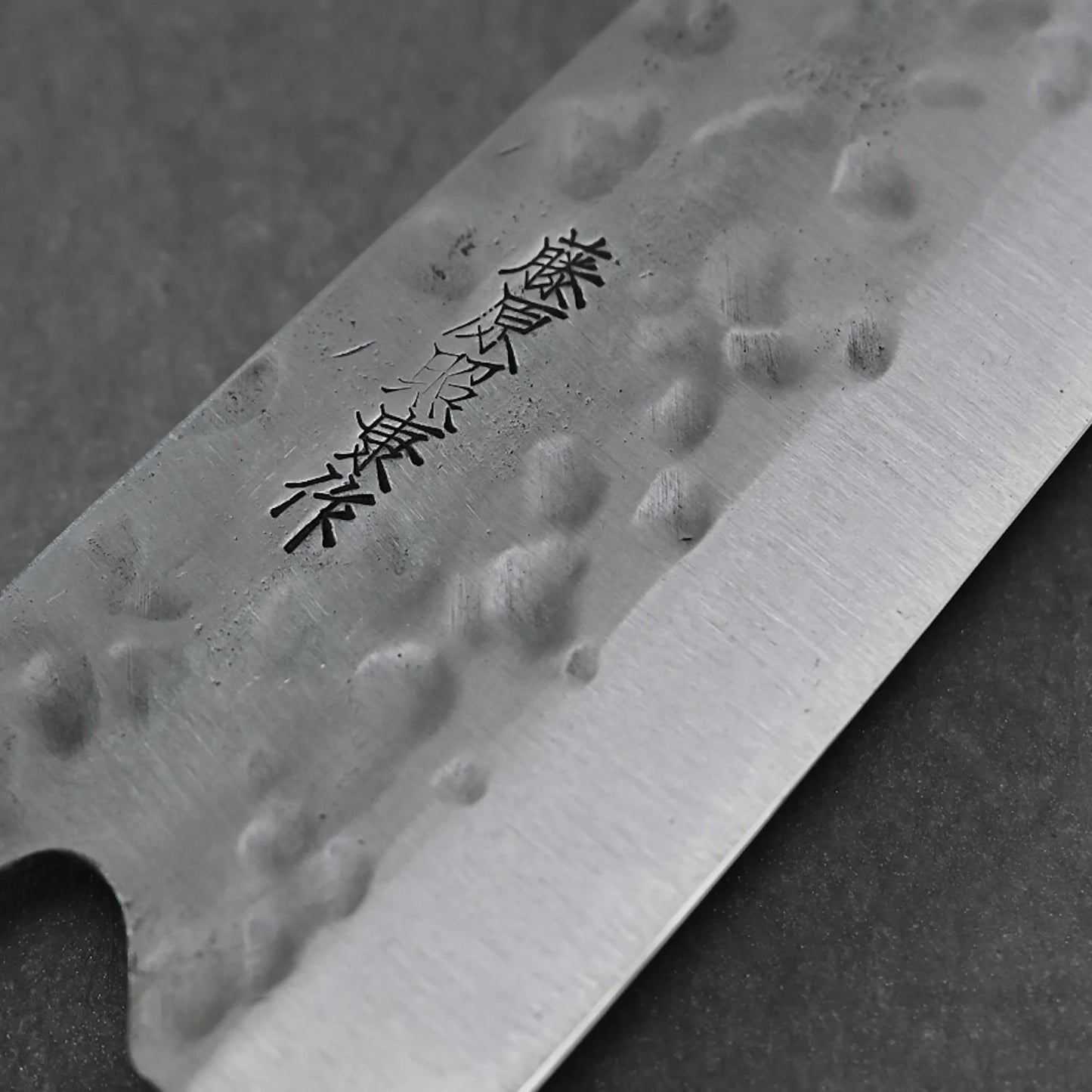 A close-up of the Teruyasu Fujiwara Maboroshi shirogami#1 wa-gyuto 210mm, featuring a hammered blade with engraved Japanese characters, custom handle, saya, and polished choil by Teruyasu Fujiwara, set against a dark background.
