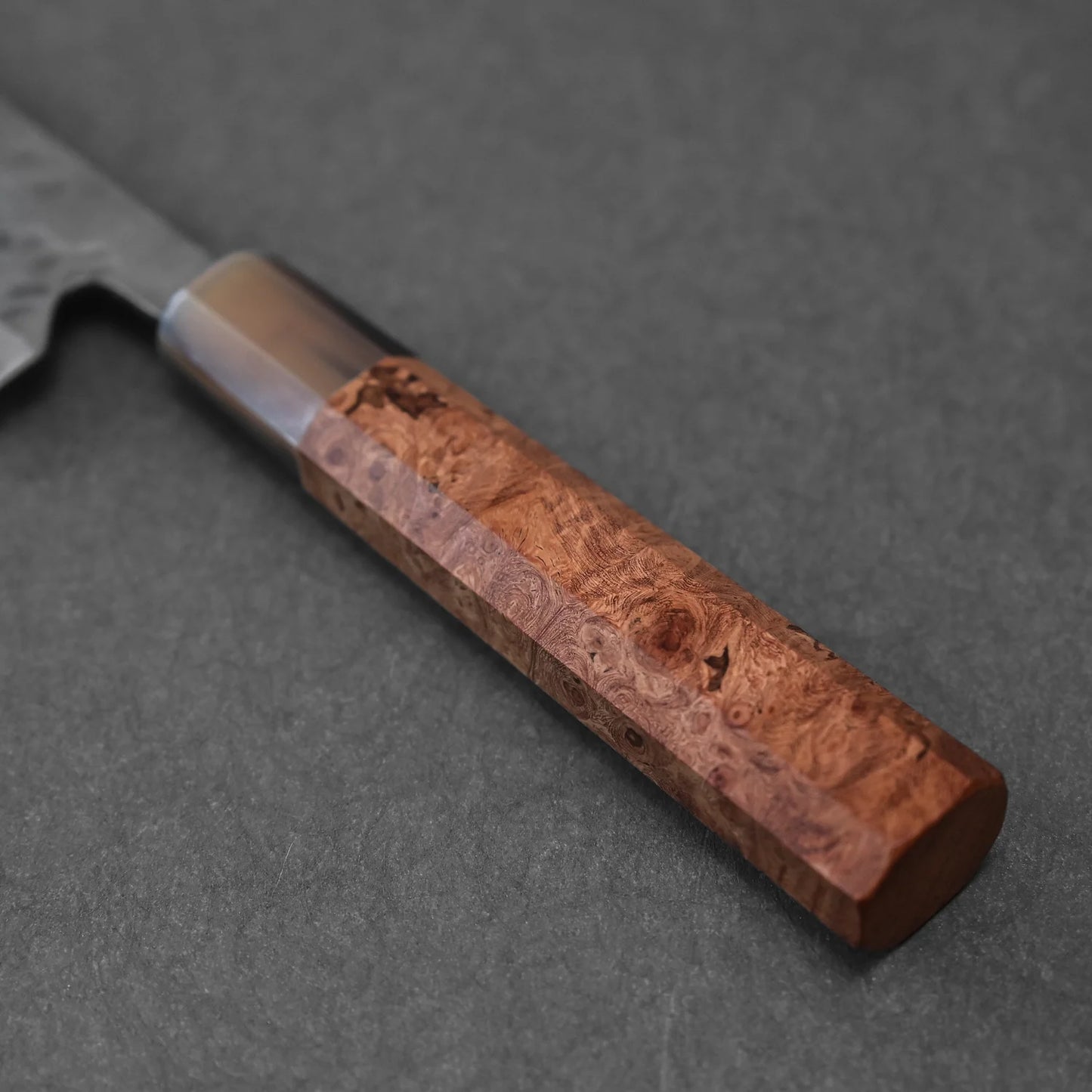 Close-up of the Teruyasu Fujiwara Maboroshi shirogami#1 wa-gyuto 210mm’s custom textured wood handle, set on a dark gray background; its polished choil and part of the blade are visible on the left.