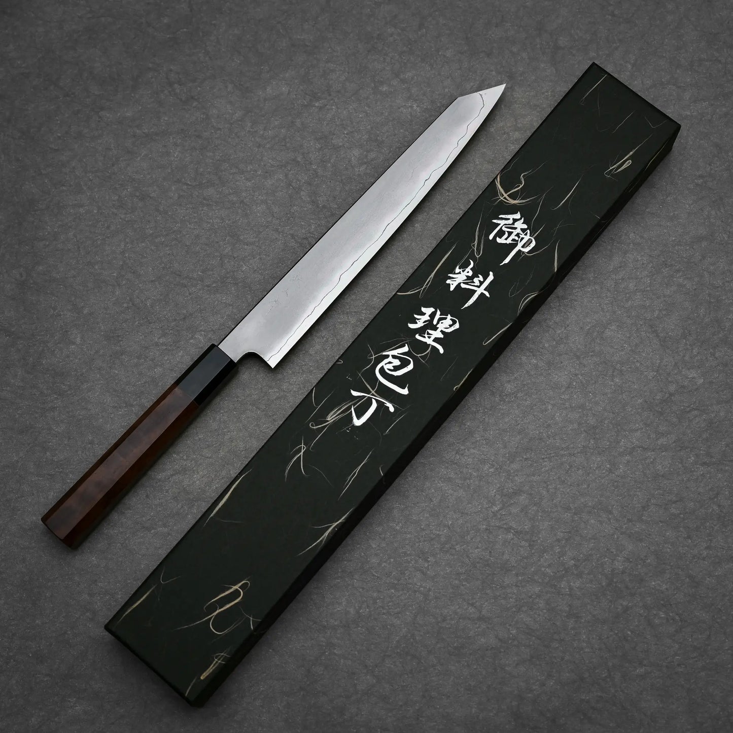 The Tetsujin kasumi ginsan kiritsuke sujihiki 240mm, featuring a rectangular wooden handle and crafted from Ginsan steel with a kasumi finish, sits beside a black box with white Japanese characters on a dark textured surface.
