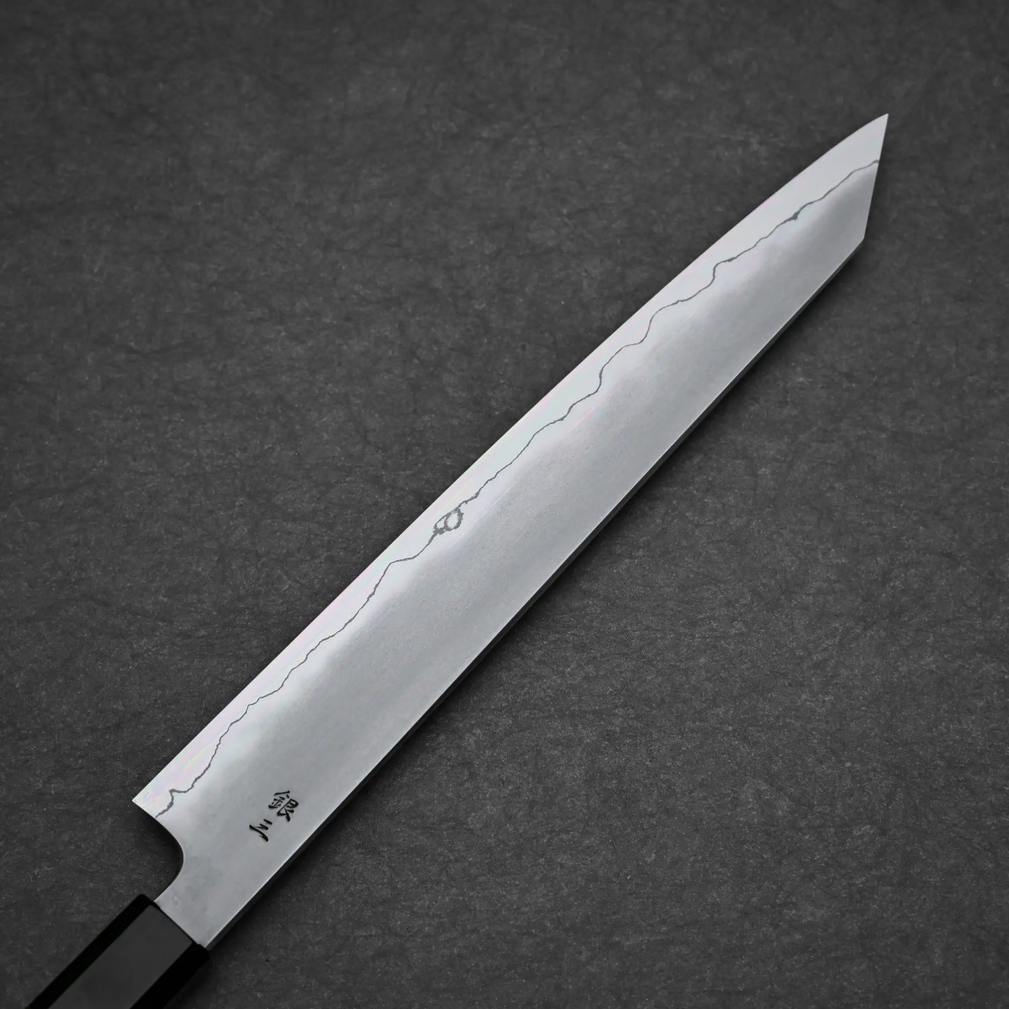 A close-up of the Tetsujin kasumi ginsan kiritsuke sujihiki 240mm knife blade, crafted by Tetsujin with Ginsan steel, highlights its wavy hamon line and subtle kasumi finish against a dark textured background.