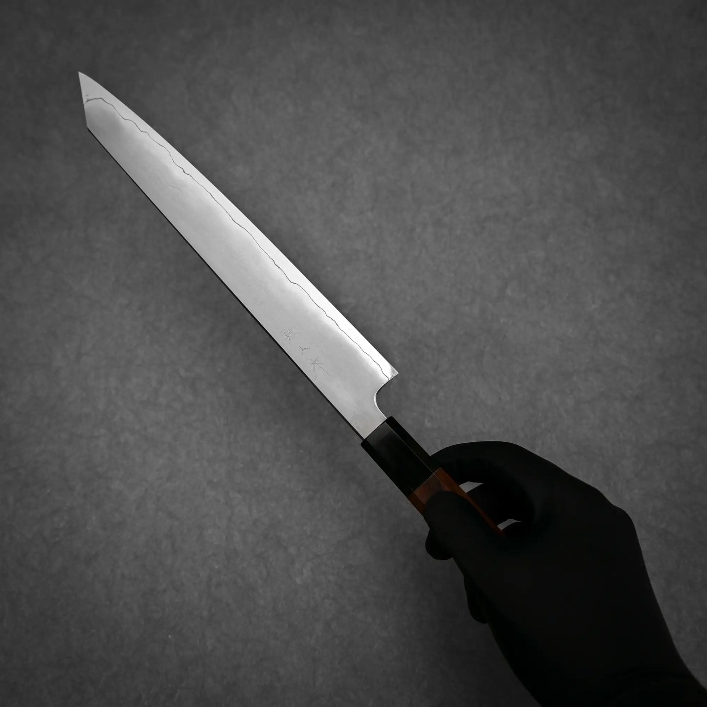 A person wearing a black glove holds the Tetsujin kasumi ginsan kiritsuke sujihiki 240mm by Tetsujin, highlighting its sharp blade and distinctive kasumi finish against a dark, textured background.