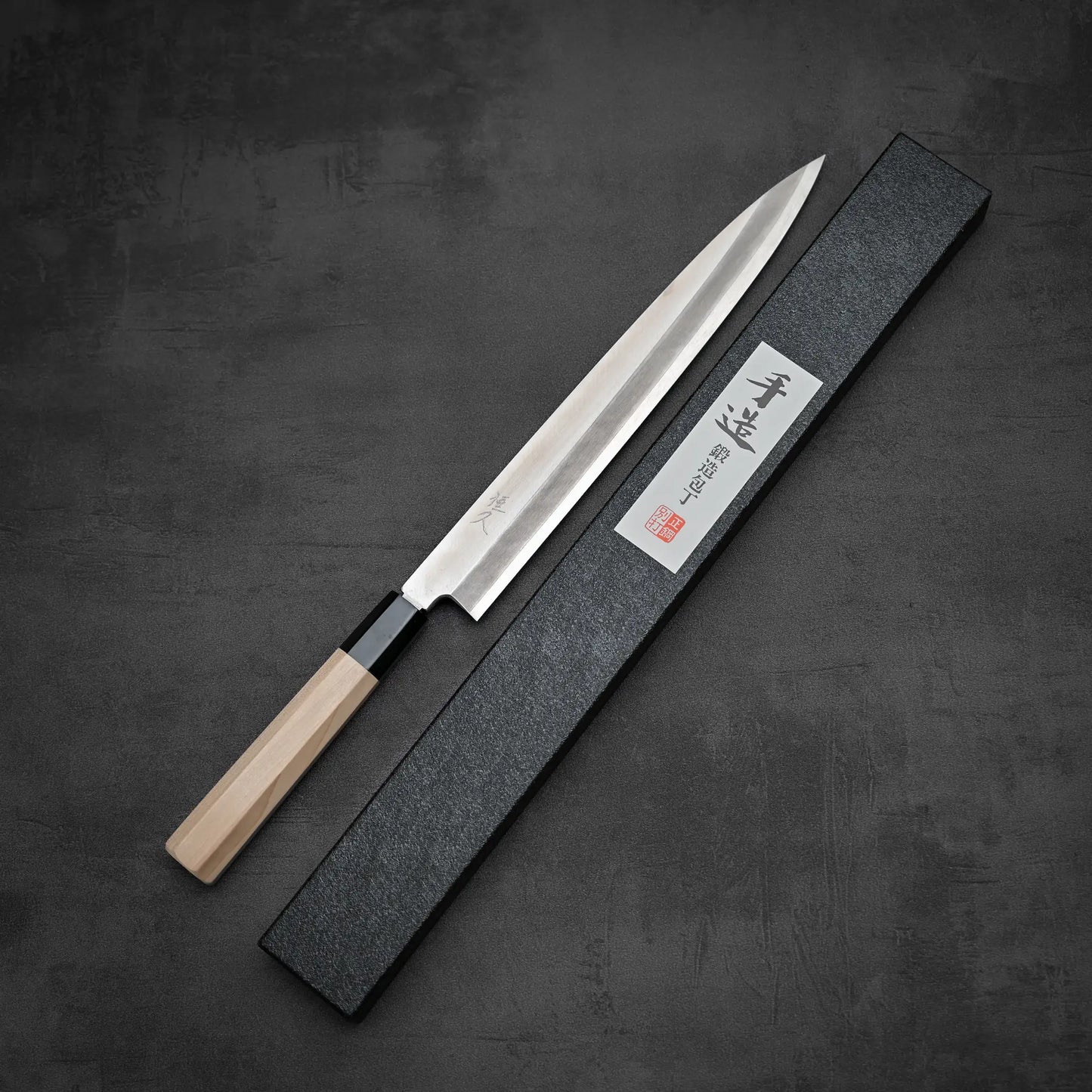 The Tsunehisa Ginsan Yanagiba 270mm, a long sashimi knife with a light wooden handle and Ginsan stainless steel blade, rests beside a black rectangular case with Japanese writing on the label atop a dark textured surface.