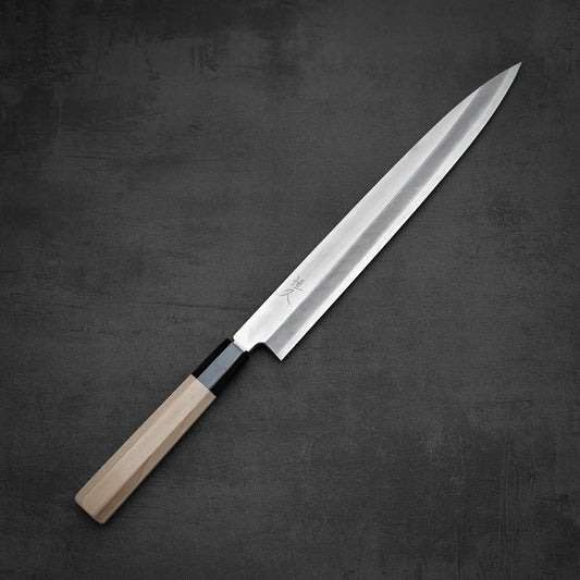 The Tsunehisa ginsan yanagiba 270mm by Tsunehisa, featuring a long Ginsan stainless steel blade and a light wood handle, rests on a dark textured surface.