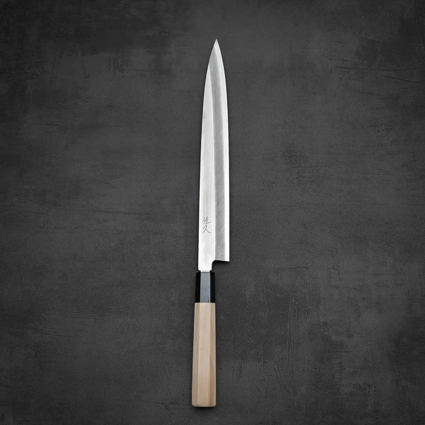 The Tsunehisa Ginsan Yanagiba 270mm, a long sashimi knife with a shiny Ginsan stainless steel blade and light wooden handle, rests on a dark, textured surface.