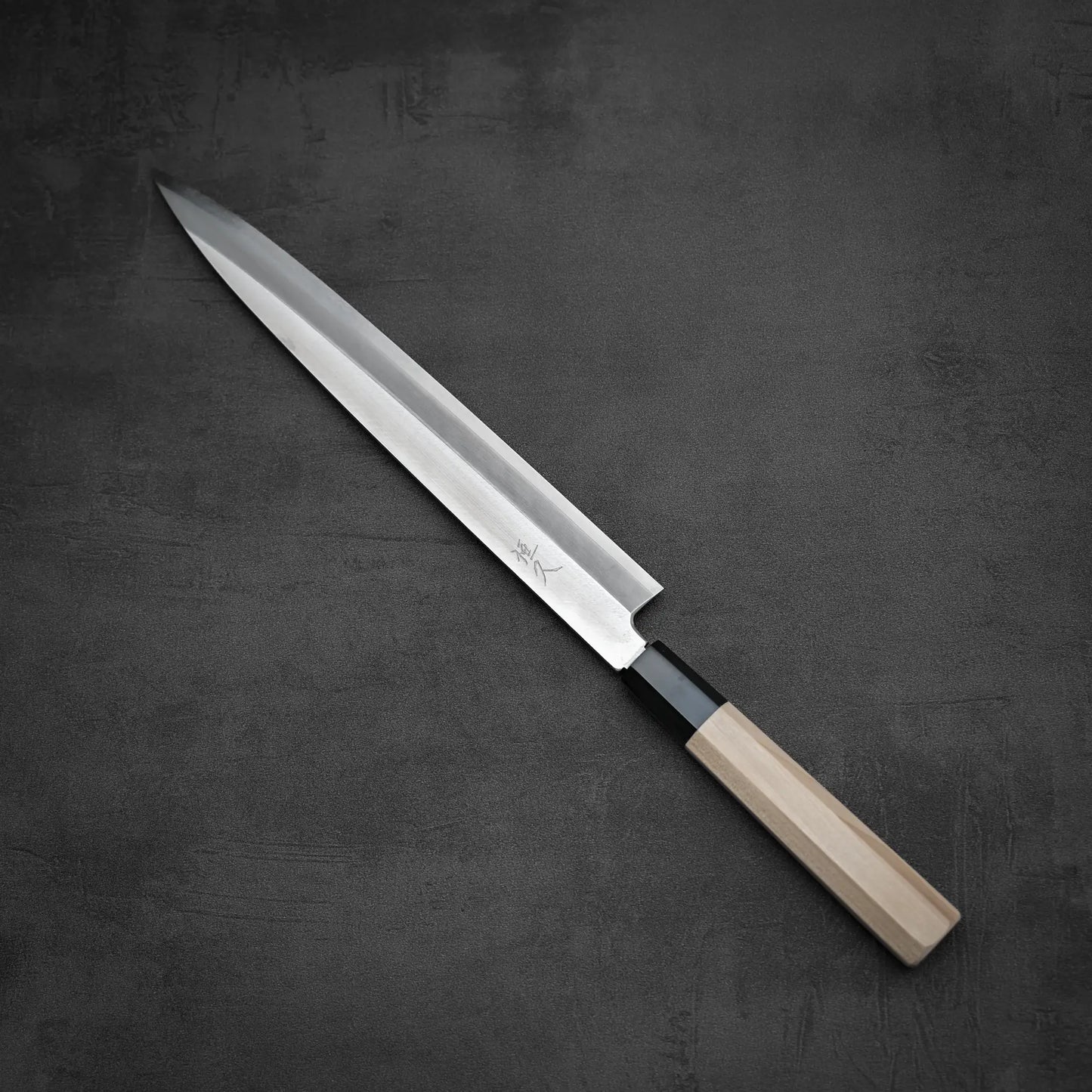 The Tsunehisa ginsan yanagiba 270mm, featuring a Ginsan stainless steel blade and a light wooden handle, is positioned diagonally on a dark gray textured surface.