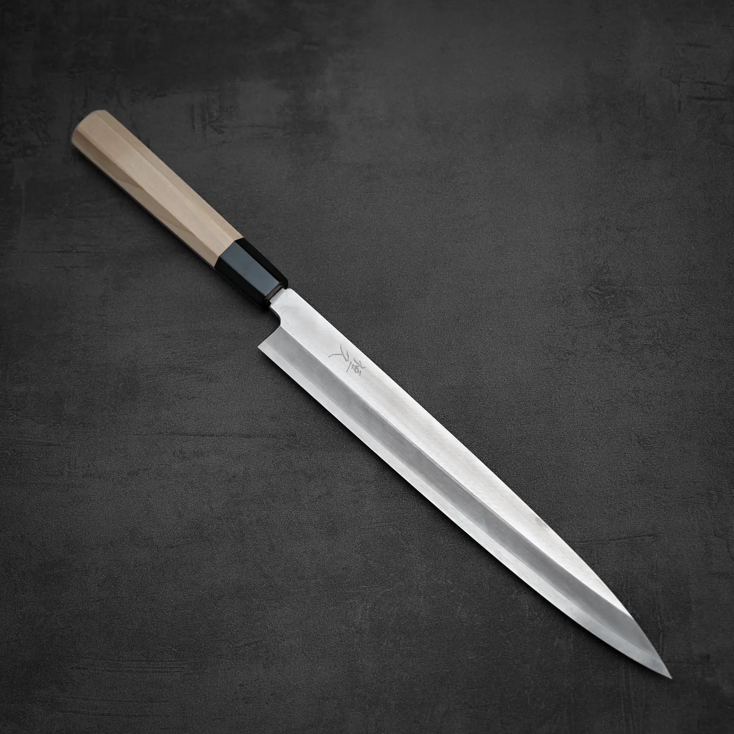 The Tsunehisa ginsan yanagiba 270mm, featuring a long Ginsan stainless steel blade and a light wooden handle, rests on a dark textured surface.