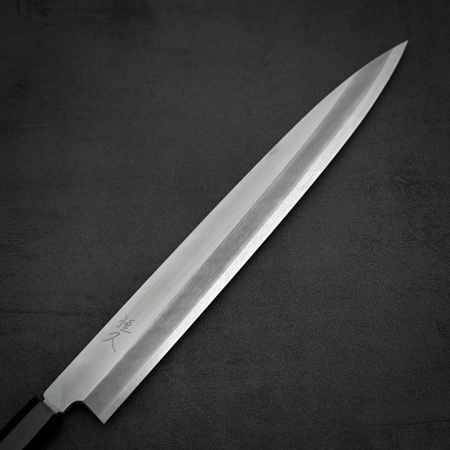 The Tsunehisa ginsan yanagiba 270mm, a Japanese-style sashimi knife with a long, narrow Ginsan stainless steel blade and black handle, bearing engraved Japanese characters near the handle, rests on a dark textured surface.