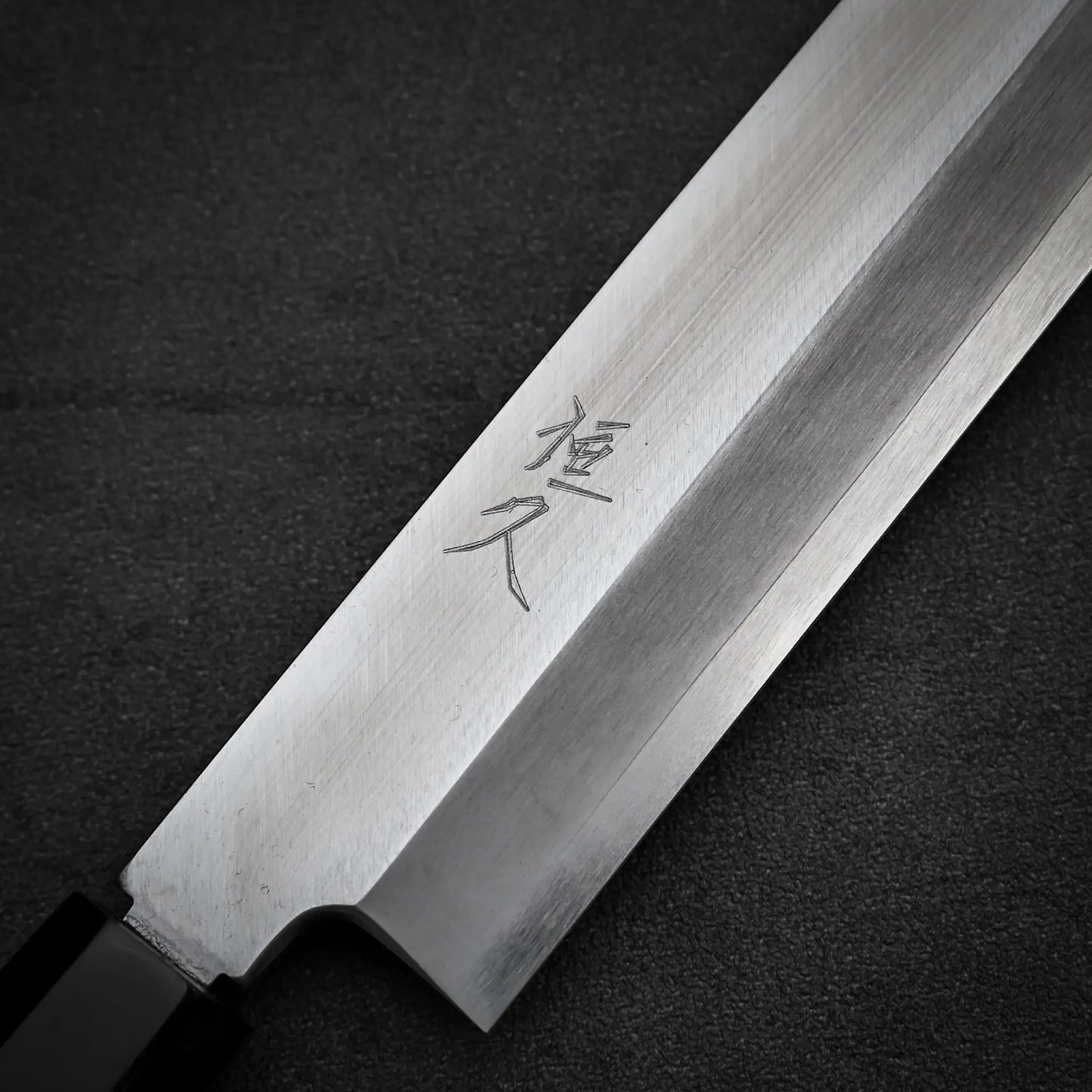 A close-up of the Tsunehisa ginsan yanagiba 270mm, a Japanese sashimi knife by Tsunehisa with kanji engraved on its Ginsan stainless steel blade, laid on a dark textured surface.