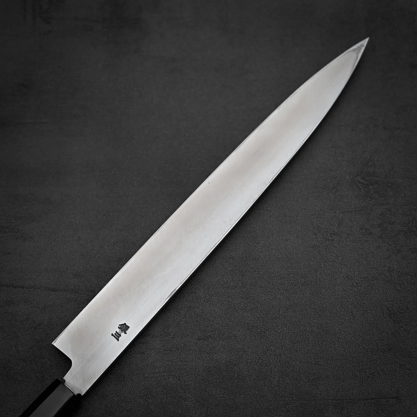 The Tsunehisa ginsan yanagiba 270mm, a long Japanese sashimi knife with a sharp Ginsan stainless steel blade and black handle, lies on a dark, textured surface.