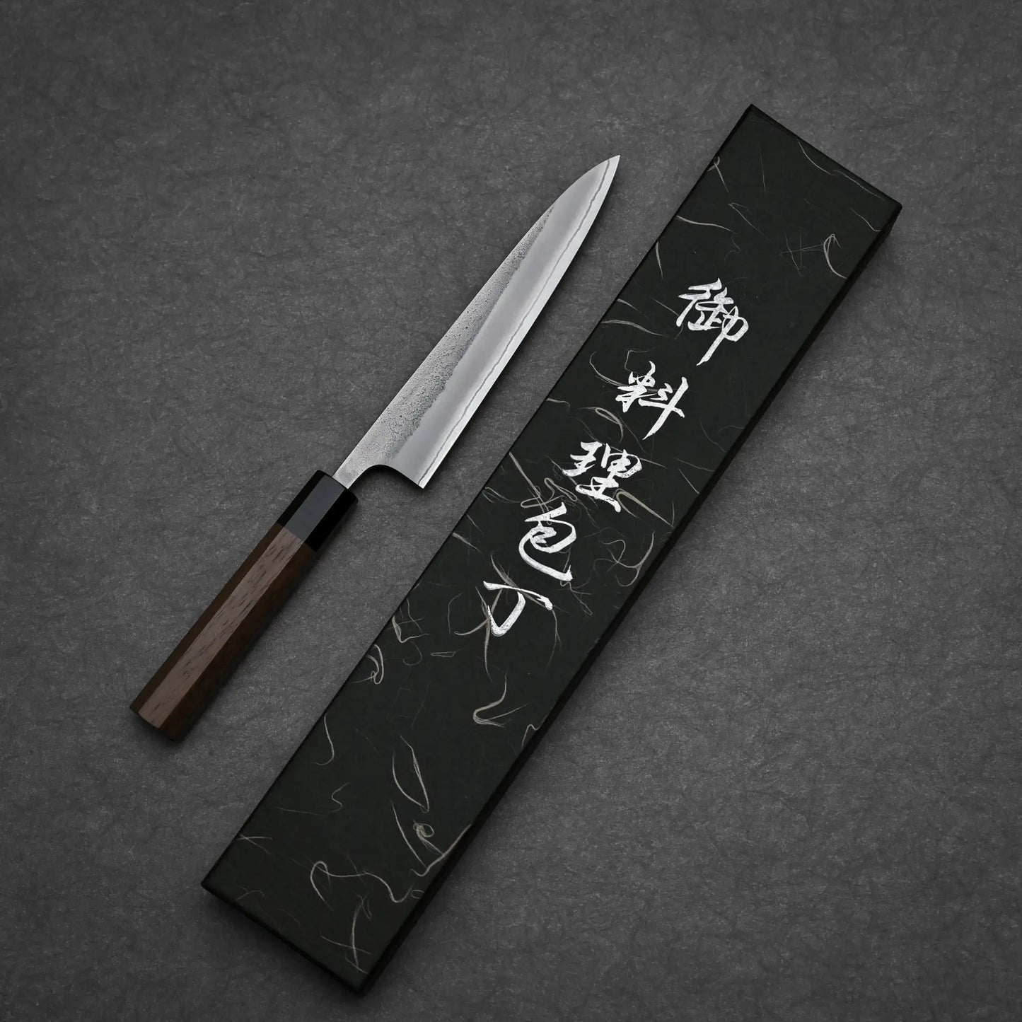 A Hatsukokoro x Yoshikane nashiji shirogami#2 petty knife 150mm with a wooden handle lies beside a rectangular black box with white Japanese characters on the lid, all set against a textured dark background.