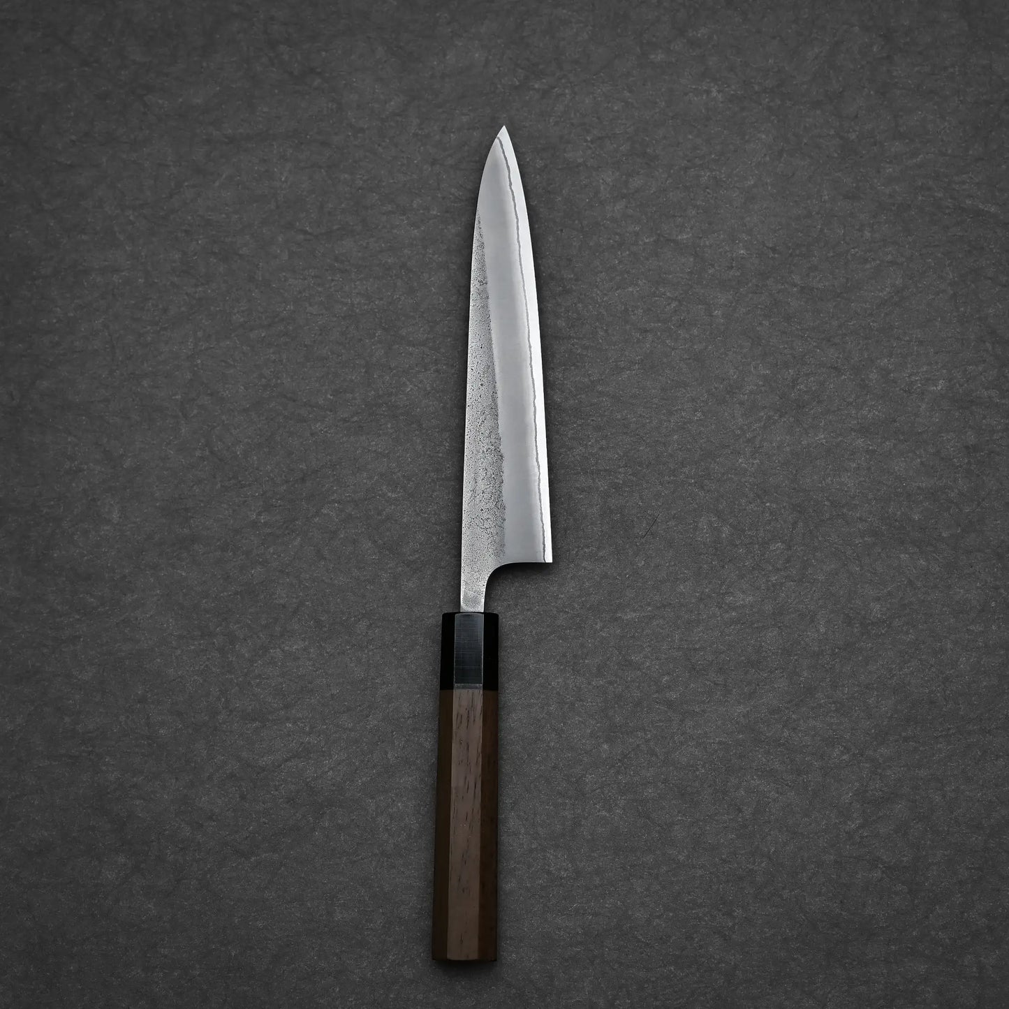 A single Yoshikane Hatsukokoro x Yoshikane nashiji shirogami#2 petty knife 150mm with a wooden handle and pointed blade rests on a dark, textured surface.