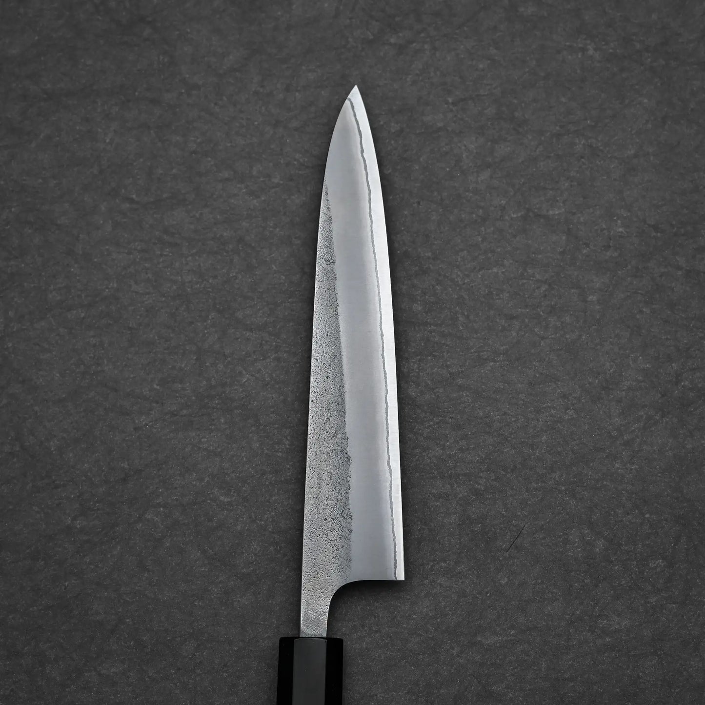 A close-up of the Yoshikane Hatsukokoro Nashiji Shirogami #2 petty knife 150mm, featuring a sharp, pointed blade and black handle, set on a dark, textured surface.