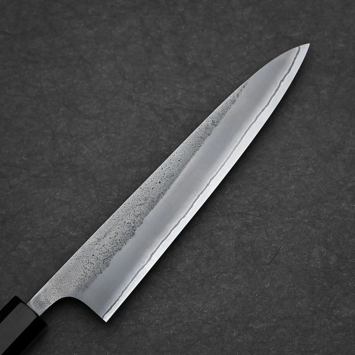 A Yoshikane Hatsukokoro nashiji shirogami#2 petty knife 150mm with a black handle lies elegantly on a dark, textured surface.