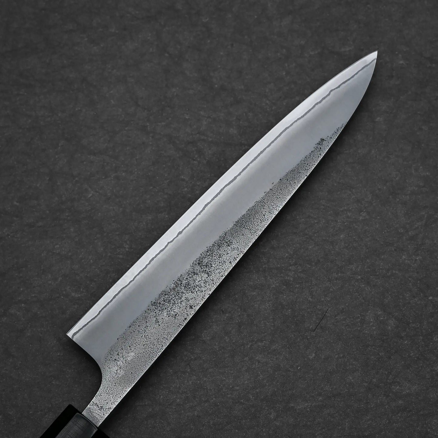 A close-up of the Yoshikane Hatsukokoro x Yoshikane nashiji shirogami#2 petty knife 150mm showcases its sharp, wavy-patterned blade and textured detail against a dark, textured background.