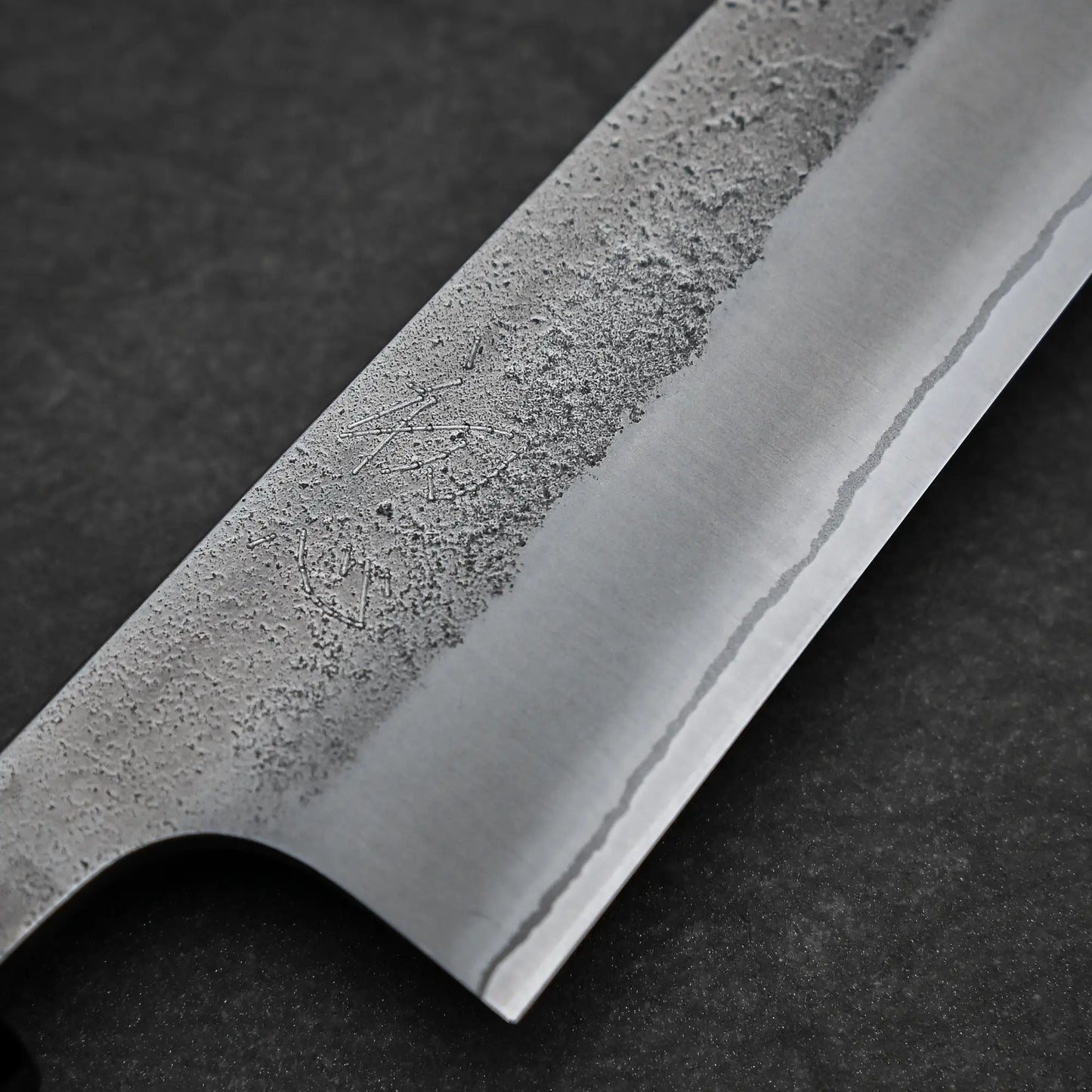 Close-up of a Yoshikane Hatsukokoro x Nashiji Shirogami #2 Petty Knife 150mm, highlighting its textured nashiji finish and polished edge, resting on a dark stone-like surface.