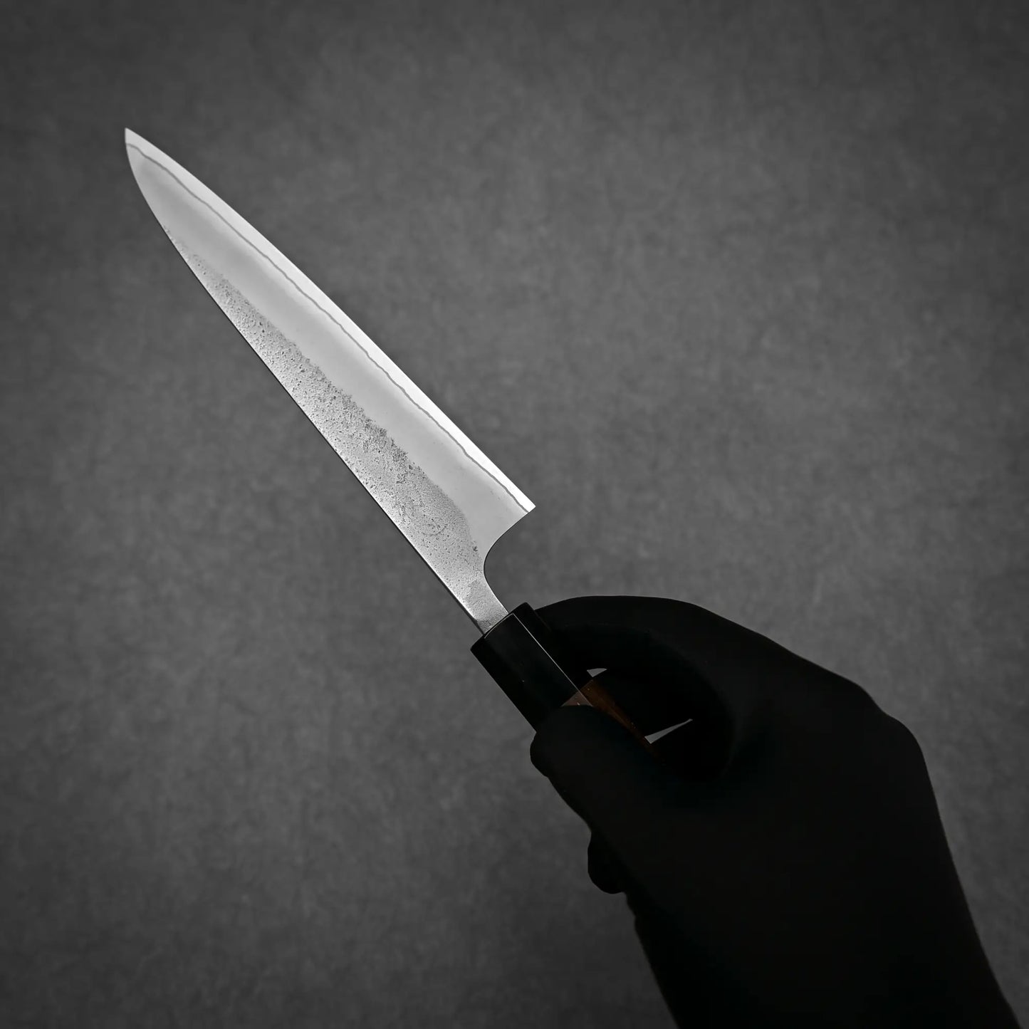 A person wearing a black glove holds a Yoshikane Hatsukokoro x Yoshikane nashiji shirogami#2 petty knife 150mm with a pointed tip and dark handle against a dark gray background.