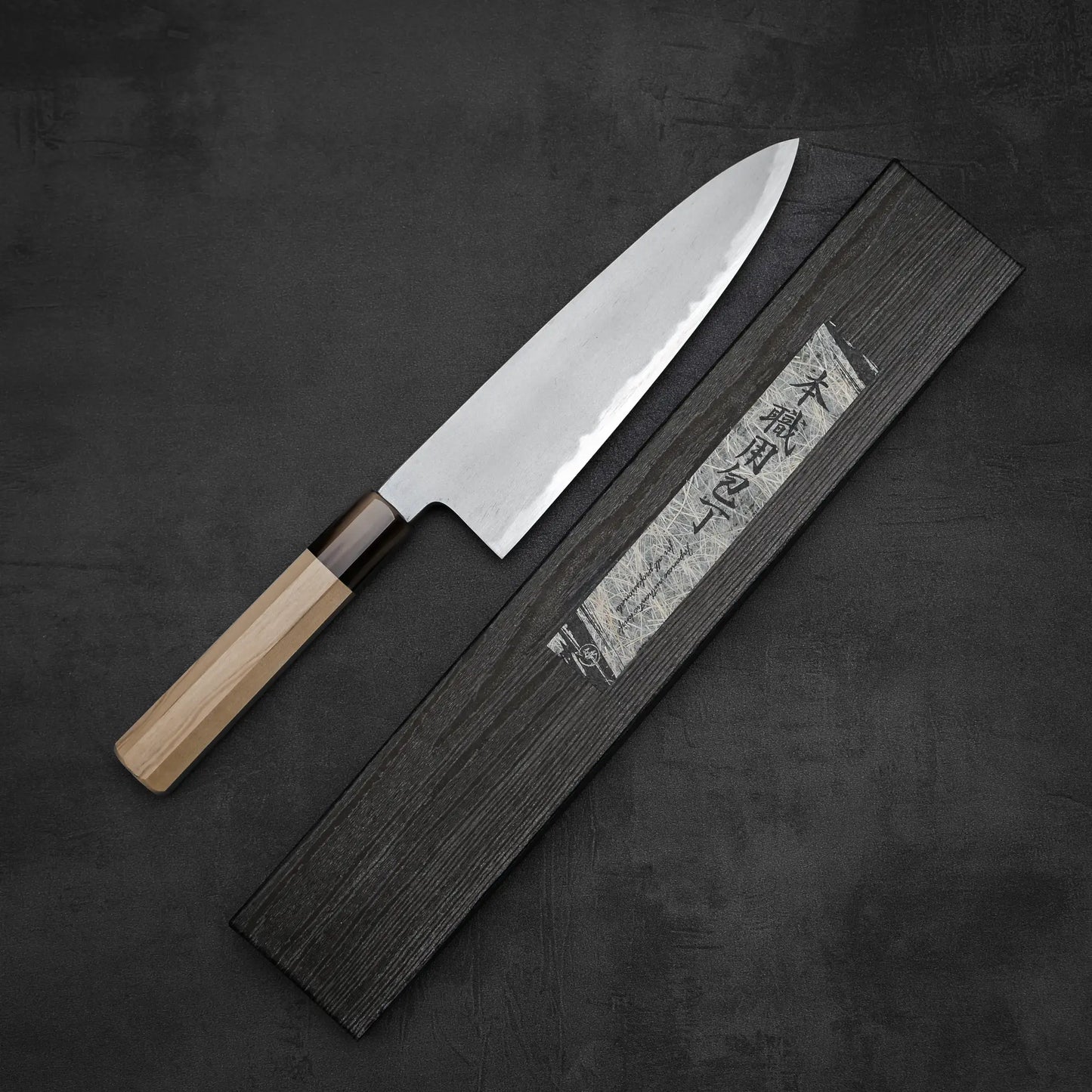 The Yoshikazu Tanaka AS tall gyuto 210mm rests diagonally on a rectangular black case with Japanese characters, set against a dark textured background.