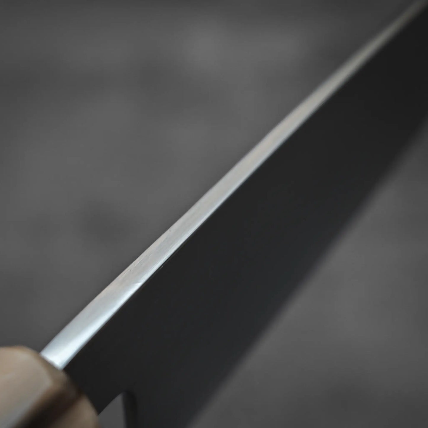 Close-up of the sharp edge of the Yoshikazu Tanaka AS tall gyuto 210mm kitchen knife by Yoshikazu Tanaka, with part of its wooden handle in view and a blurred gray background.