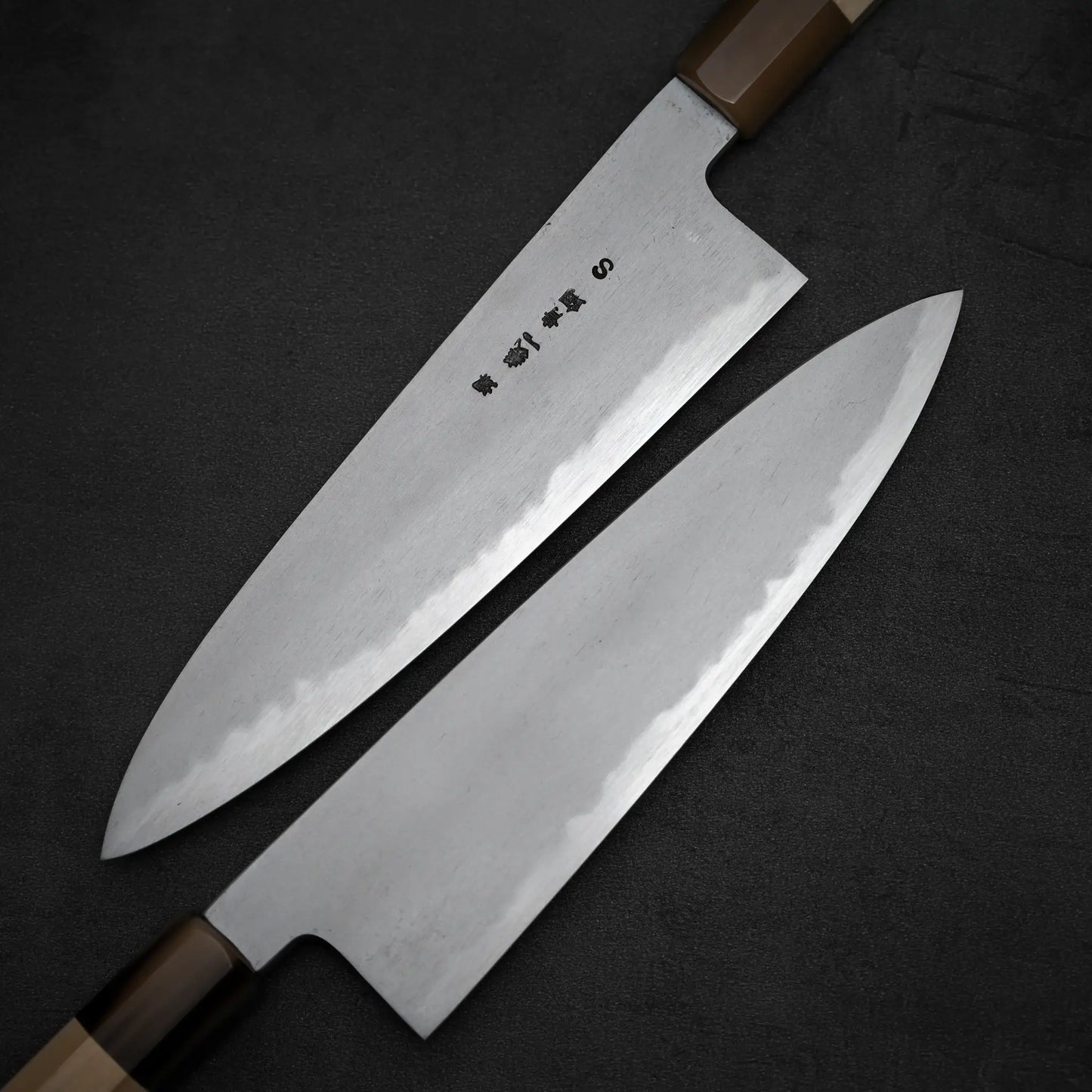 Two Japanese kitchen knives with wooden handles sit on a dark surface: one, the Yoshikazu Tanaka AS tall gyuto 210mm, features engraved characters and a sharp Aogami Super steel blade; the other, unadorned, shows off its keen edge.