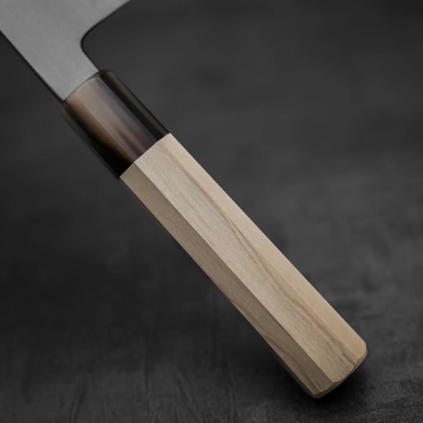 Close-up of a Yoshikazu Tanaka AS tall gyuto 210mm kitchen knife handle with light wood and a dark bolster, crafted from Aogami Super steel by Yoshikazu Tanaka, set against a dark, blurred background.