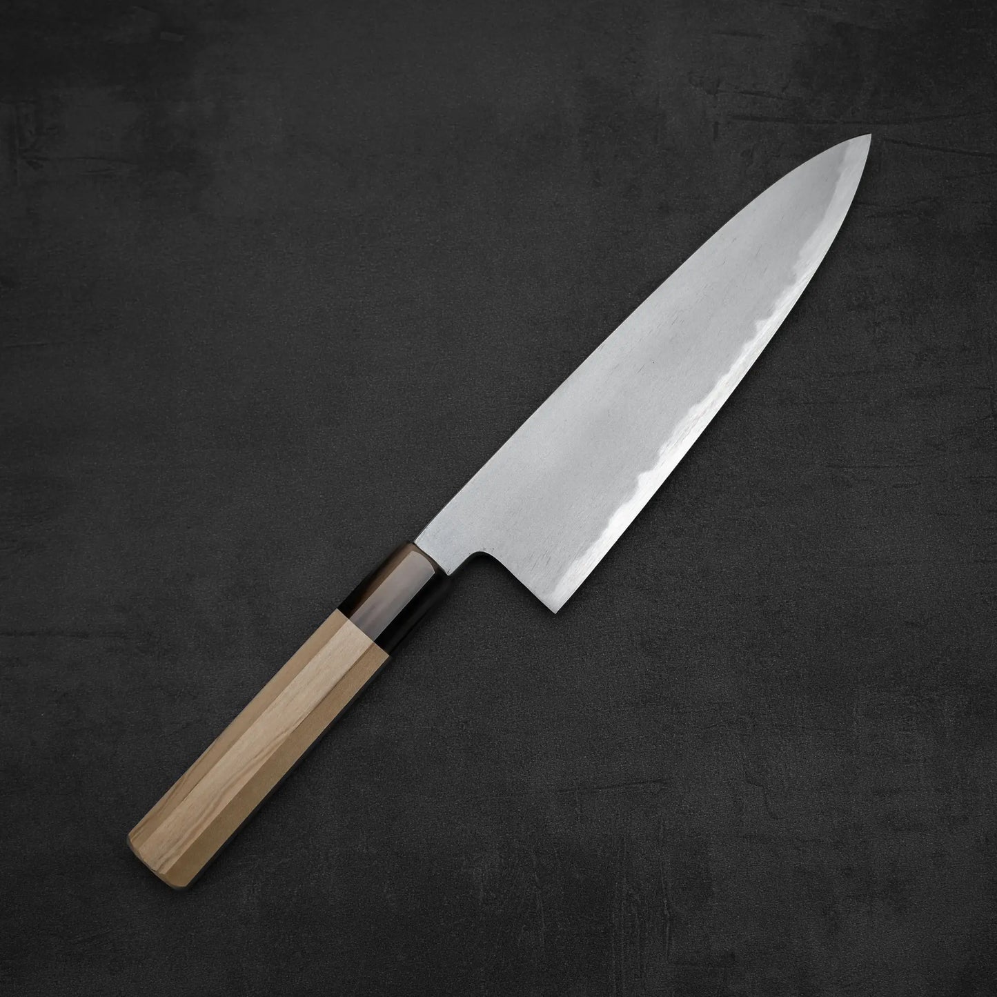 A Yoshikazu Tanaka AS tall gyuto 210mm chef’s knife with a light wood handle and sharp Aogami Super steel blade from Yoshikazu Tanaka is placed on a dark, textured surface.