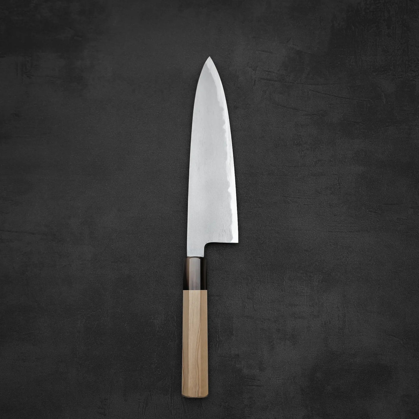 The Yoshikazu Tanaka AS tall gyuto 210mm features a wooden handle and a polished, pointed blade, showcased elegantly on a dark, textured surface.