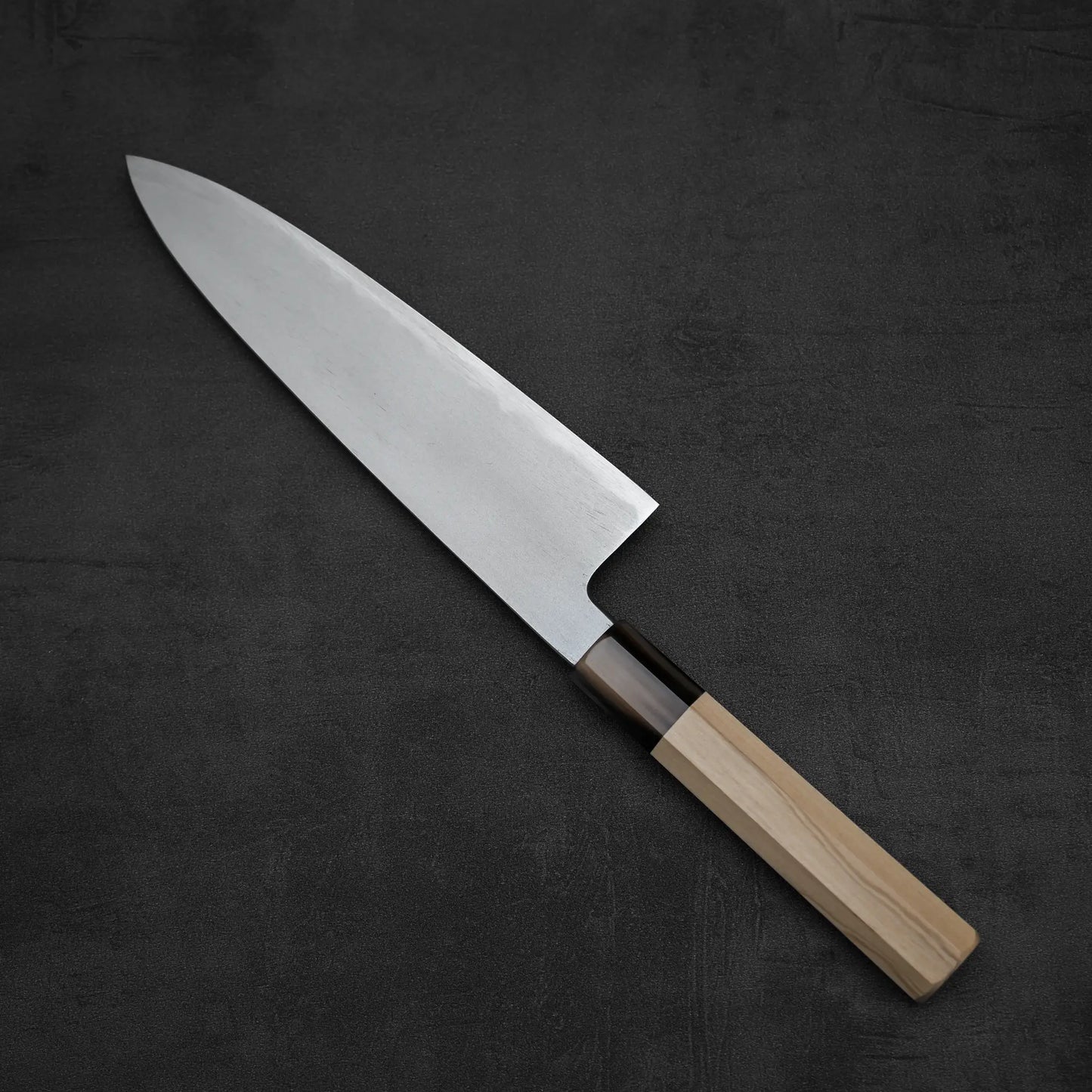 A Yoshikazu Tanaka AS tall gyuto 210mm chef’s knife with a wide stainless steel blade and a light wooden handle is placed on a dark, textured surface.