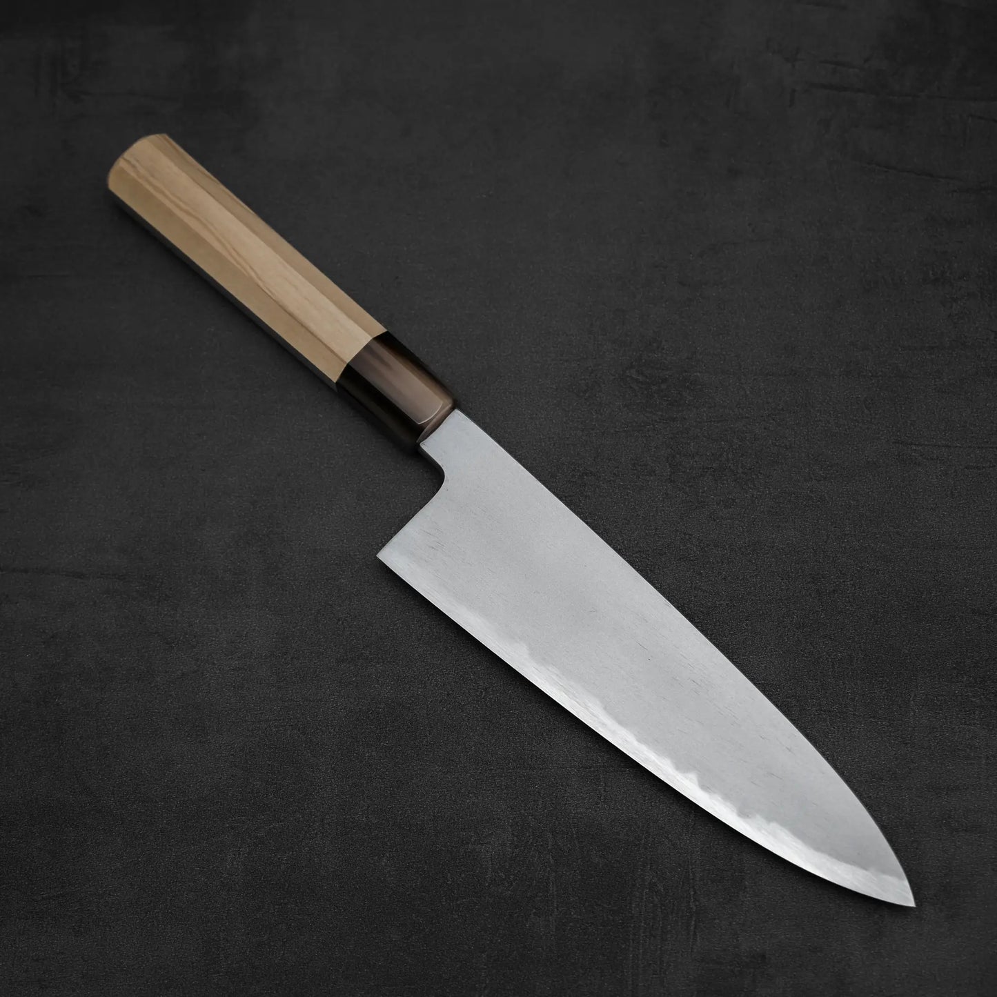 A Yoshikazu Tanaka AS tall gyuto 210mm chef’s knife by Yoshikazu Tanaka rests diagonally on a dark, textured surface.
