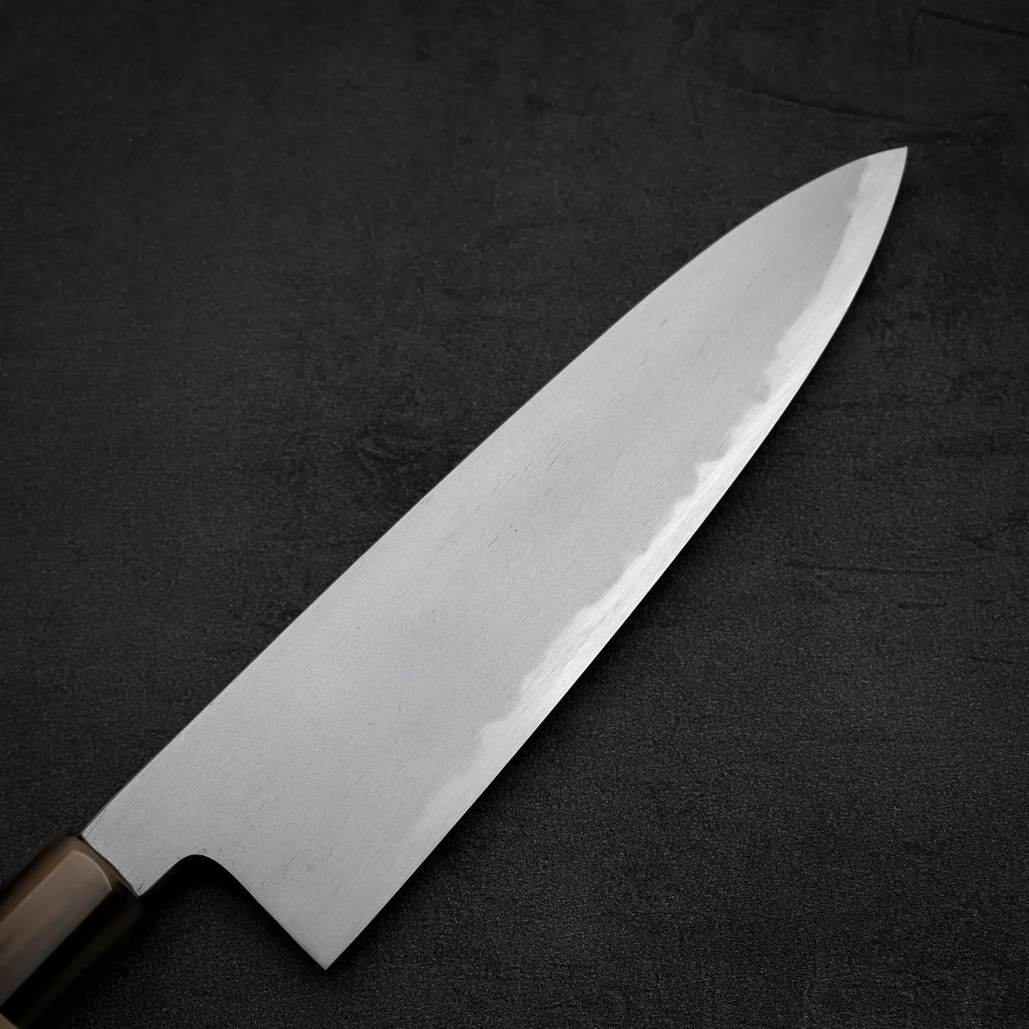 A close-up of the Yoshikazu Tanaka AS tall gyuto 210mm chef’s knife, showcasing its sharp, shiny blade and dark handle, crafted by Yoshikazu Tanaka and set against a plain, dark textured background.
