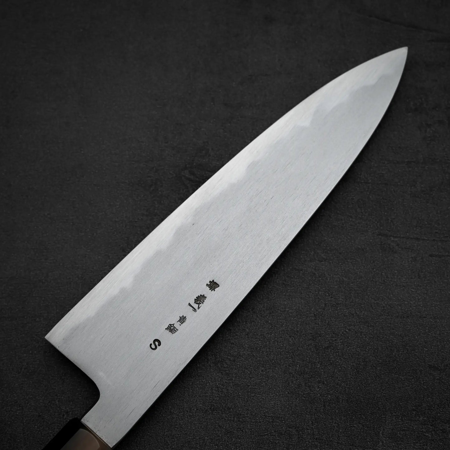 The Yoshikazu Tanaka AS tall gyuto 210mm, featuring an Aogami Super steel blade with kanji etching and a dark handle, is displayed on a textured dark gray surface.