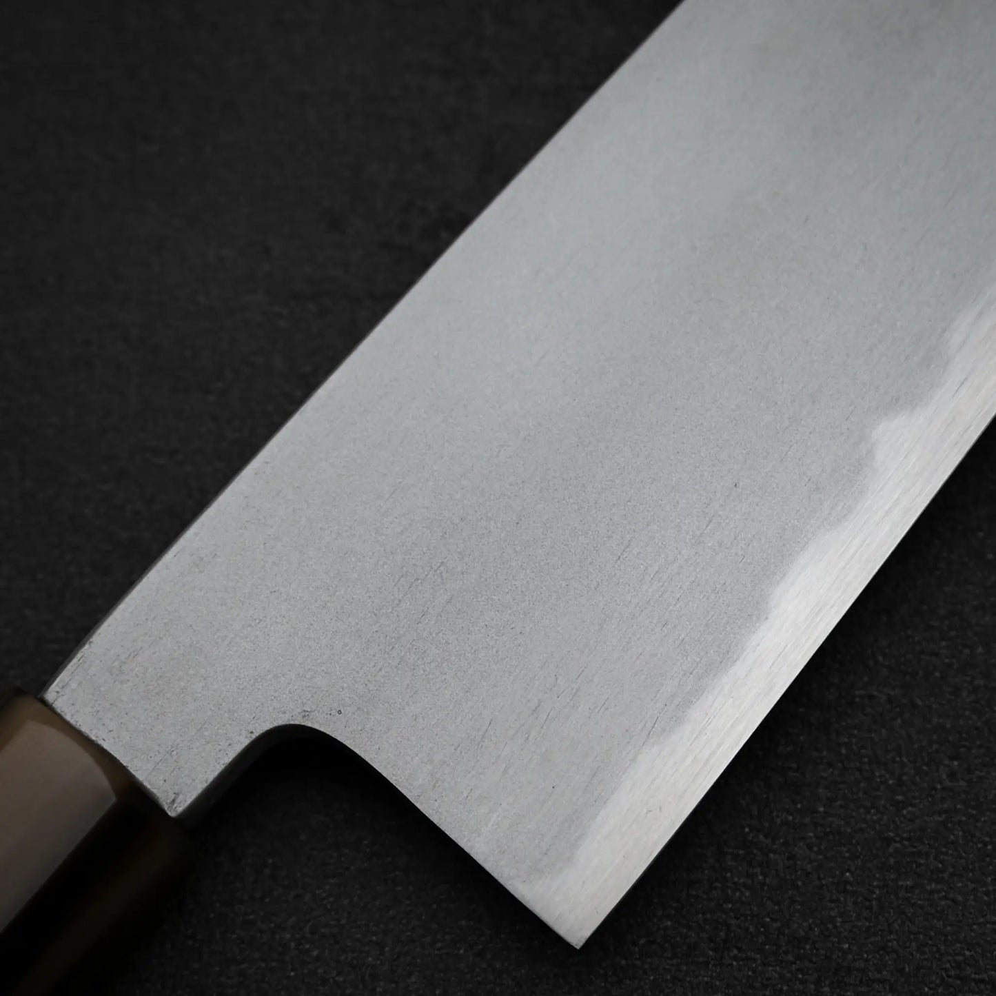 Close-up of the Yoshikazu Tanaka AS tall gyuto 210mm, showcasing its Aogami Super steel blade with a subtle pattern, polished finish, straight edge, and wooden handle set against a dark textured surface.